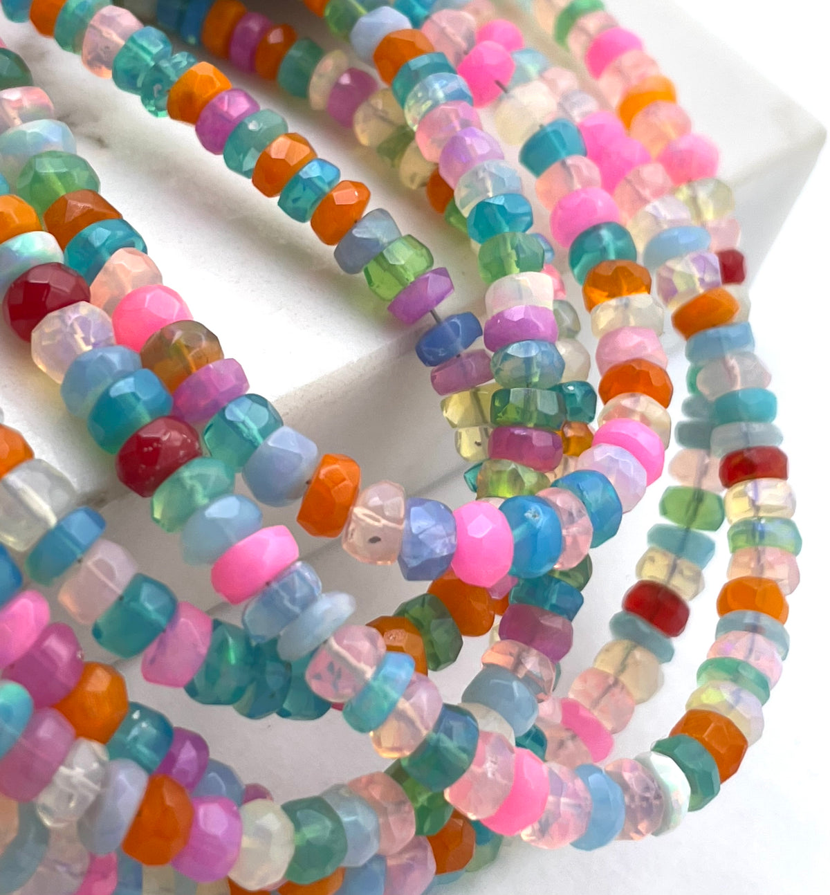 Multiple strands of faceted disc ethopian opal gemstone beads in assorted pastel and bright colors, including pink, blue, green, orange, purple, and clear, arranged on a white surface.