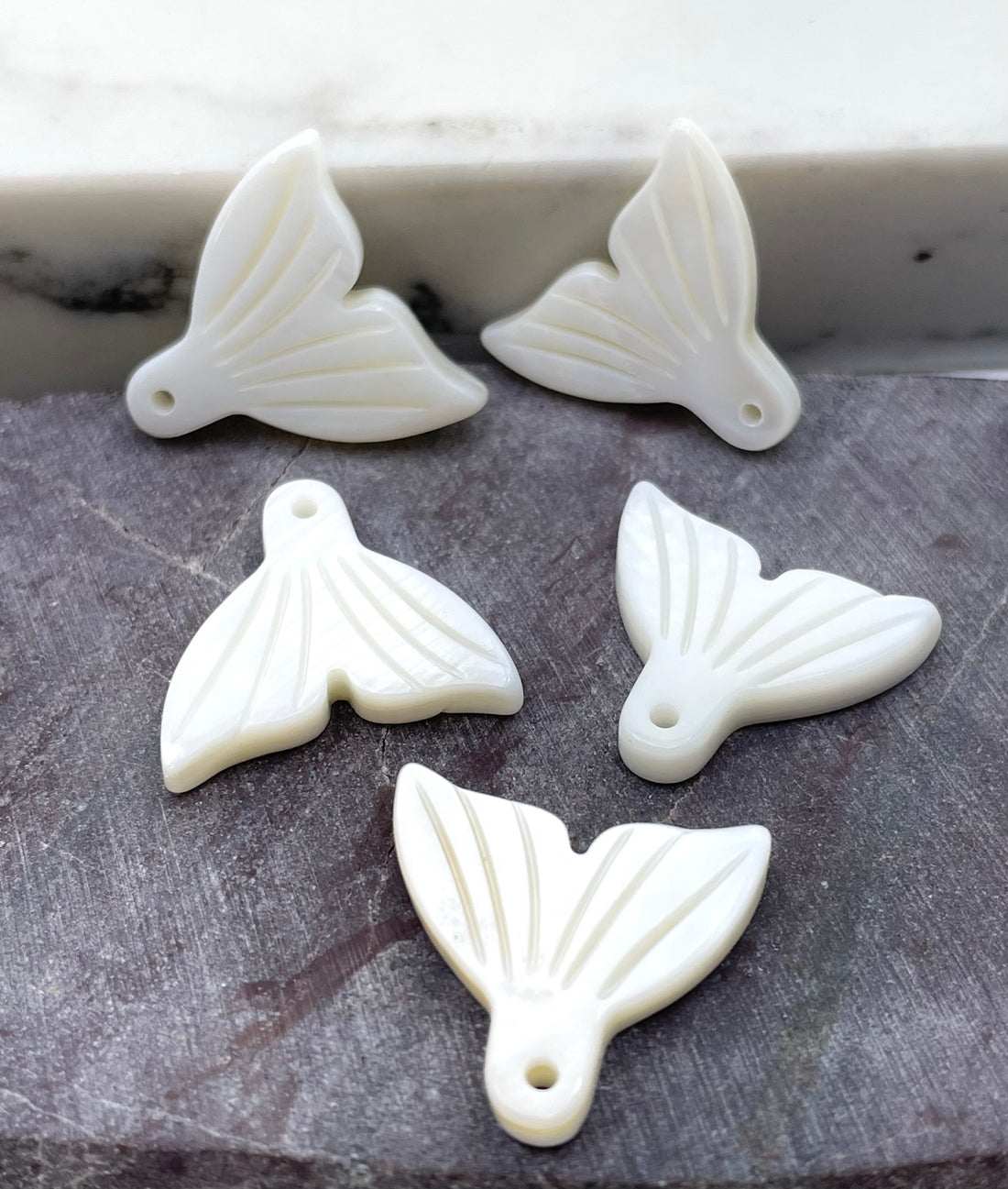 Four white carved pendant charms shaped like stylized fish or fin-like forms with a small hole, arranged on a dark textured surface.