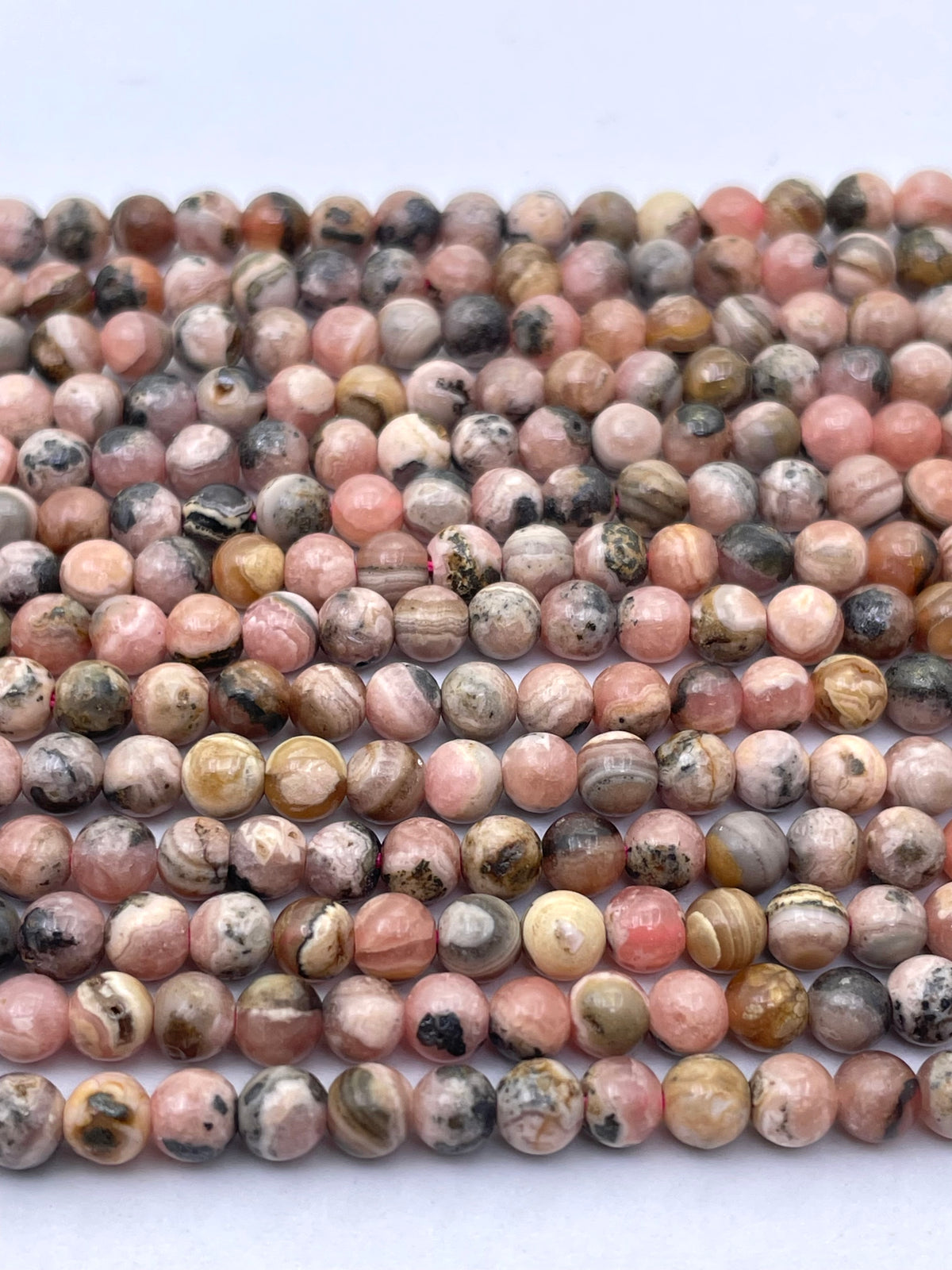 Multiple strands of round, polished gemstone beads in a marbled mix of pink, beige, brown, gray, and black, arranged horizontally on a plain white background.