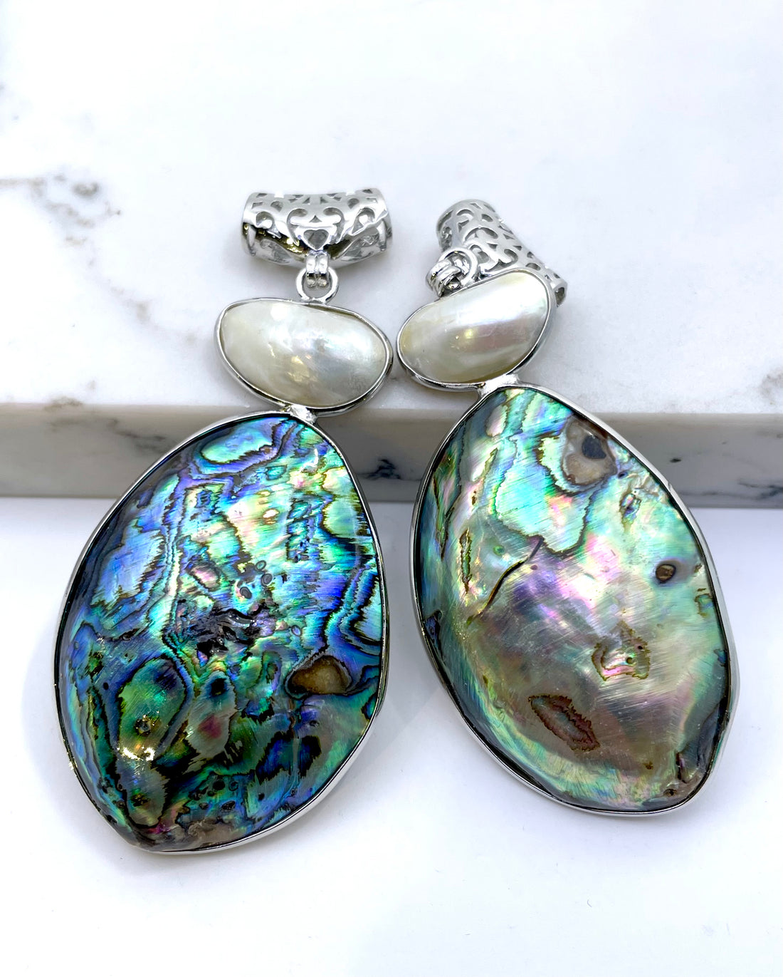 Teardrop-shaped abalone shell pendant inlays with iridescent blue, green, and purple hues, accented by small white pearls and textured silver posts, displayed on a white marble surface.