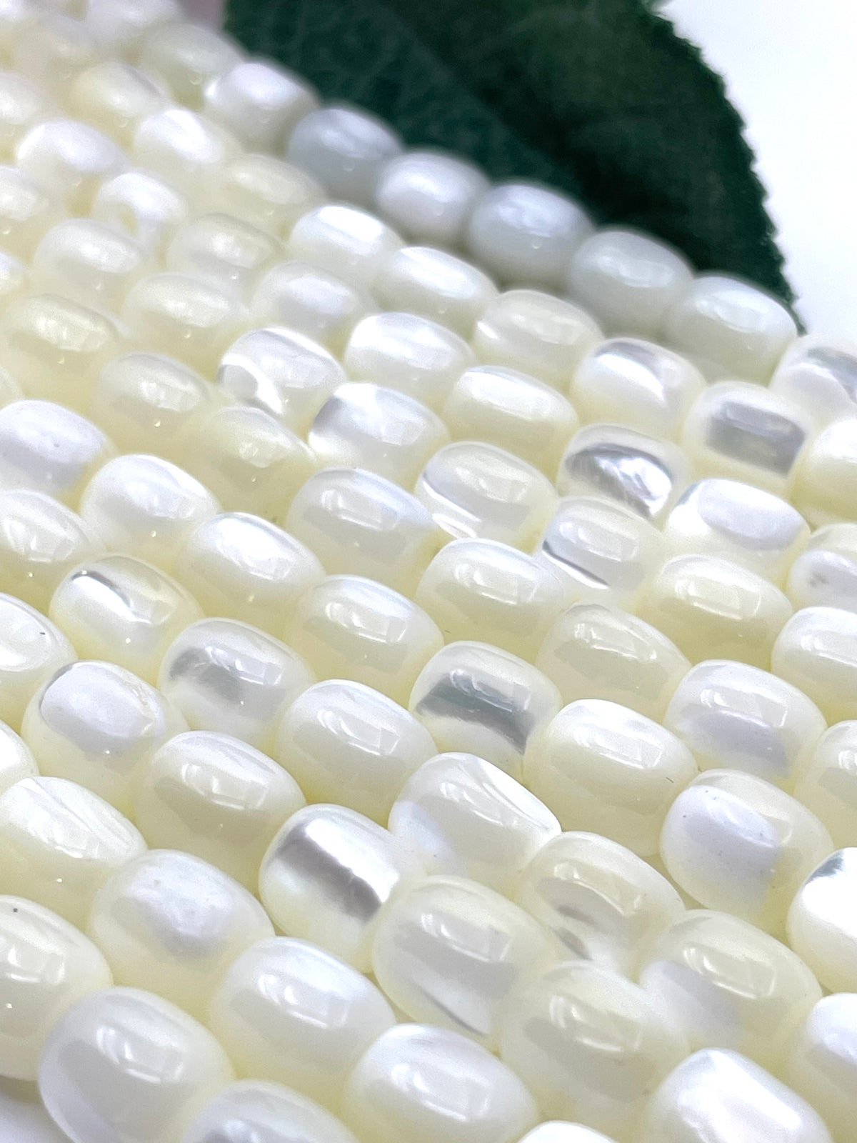 Close-up of glossy white mop oval beads arranged diagonally, with a dark green leaf partially visible in the background.