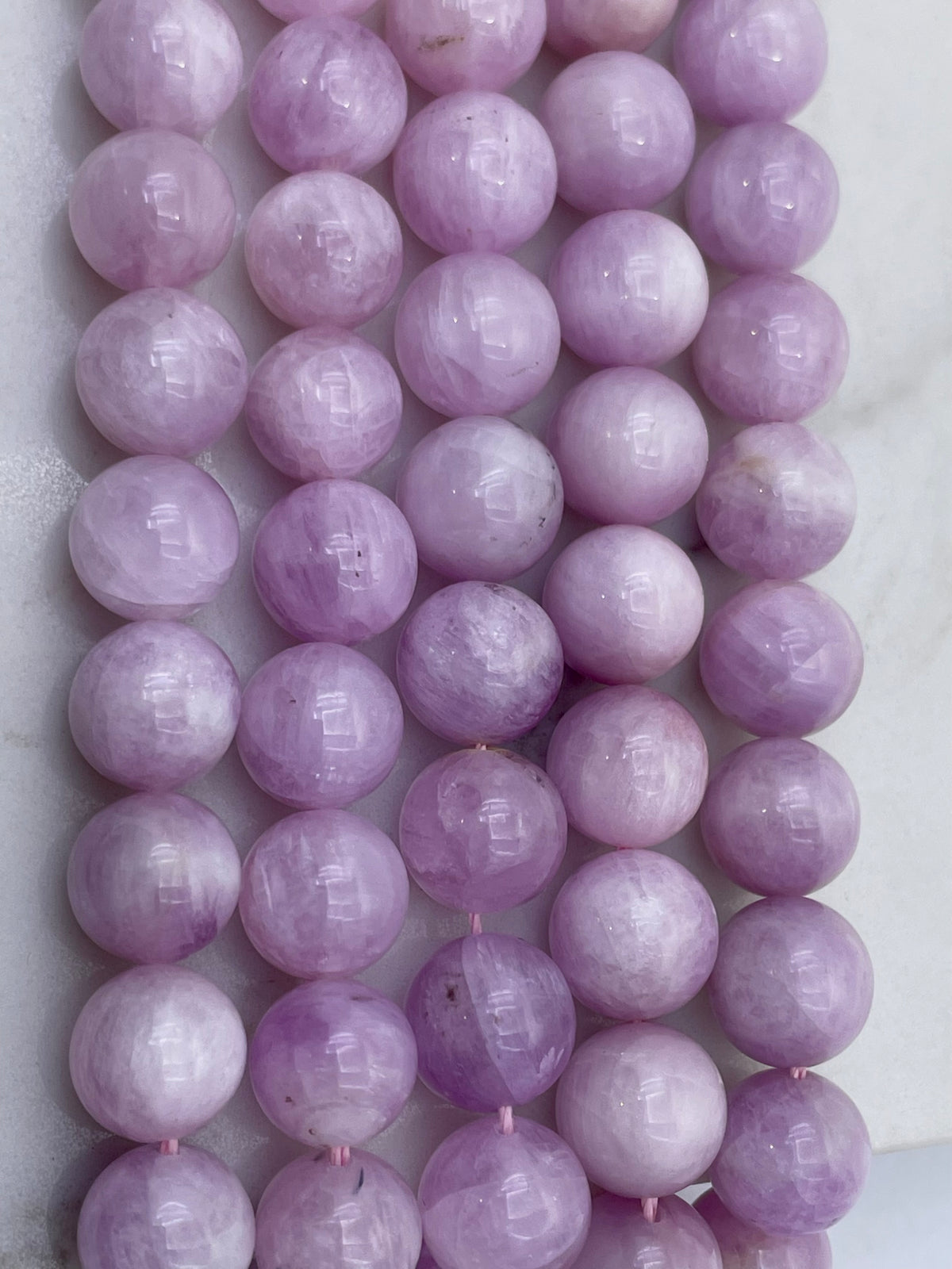 A close-up of multiple strands of polished lavender-purple kunzite gemstone beads, likely quartz or amethyst, arranged vertically on a light marble-like surface.