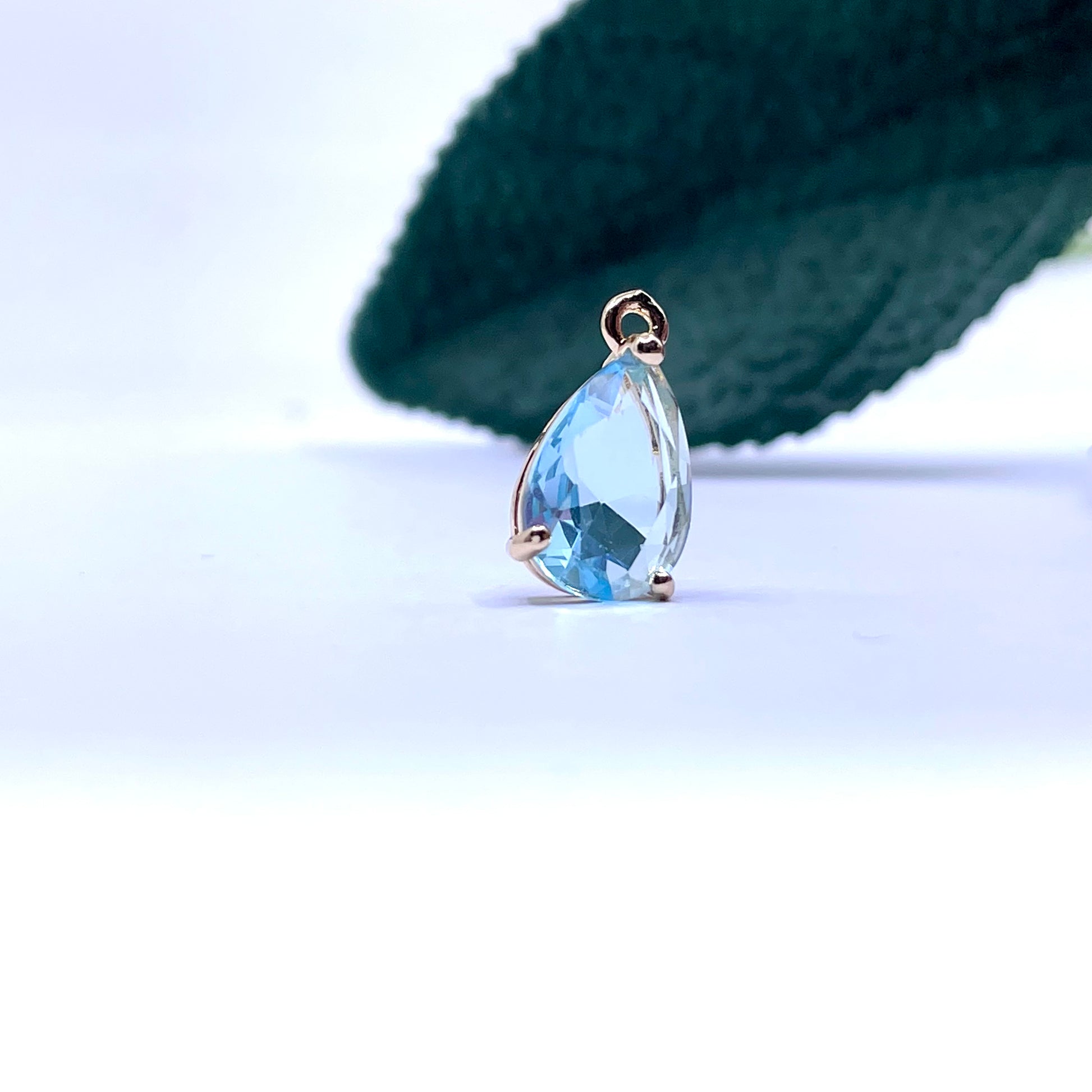 A teardrop-shaped light blue cz pendant in a gold setting, displayed on a white surface with a dark green textured background.