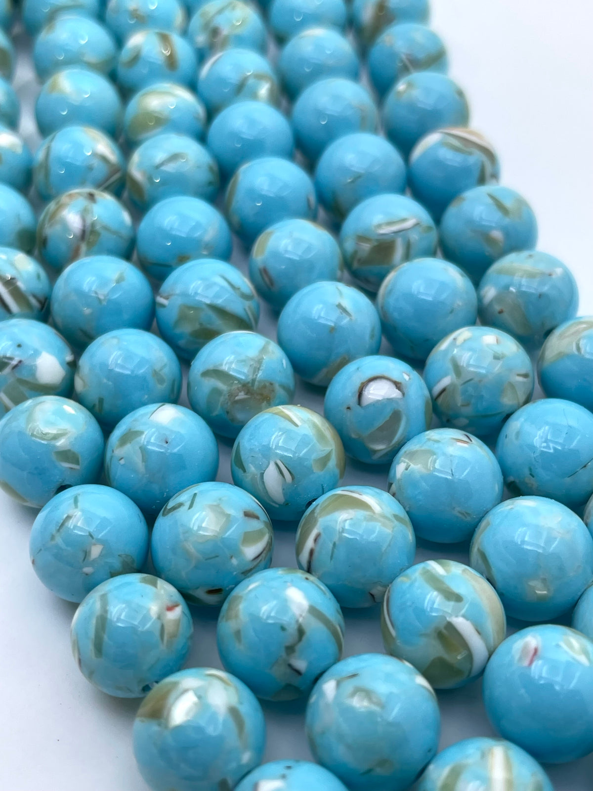 A close-up of glossy round turquoise mop beads with white and light brown marbled patterns, arranged on a plain white background.