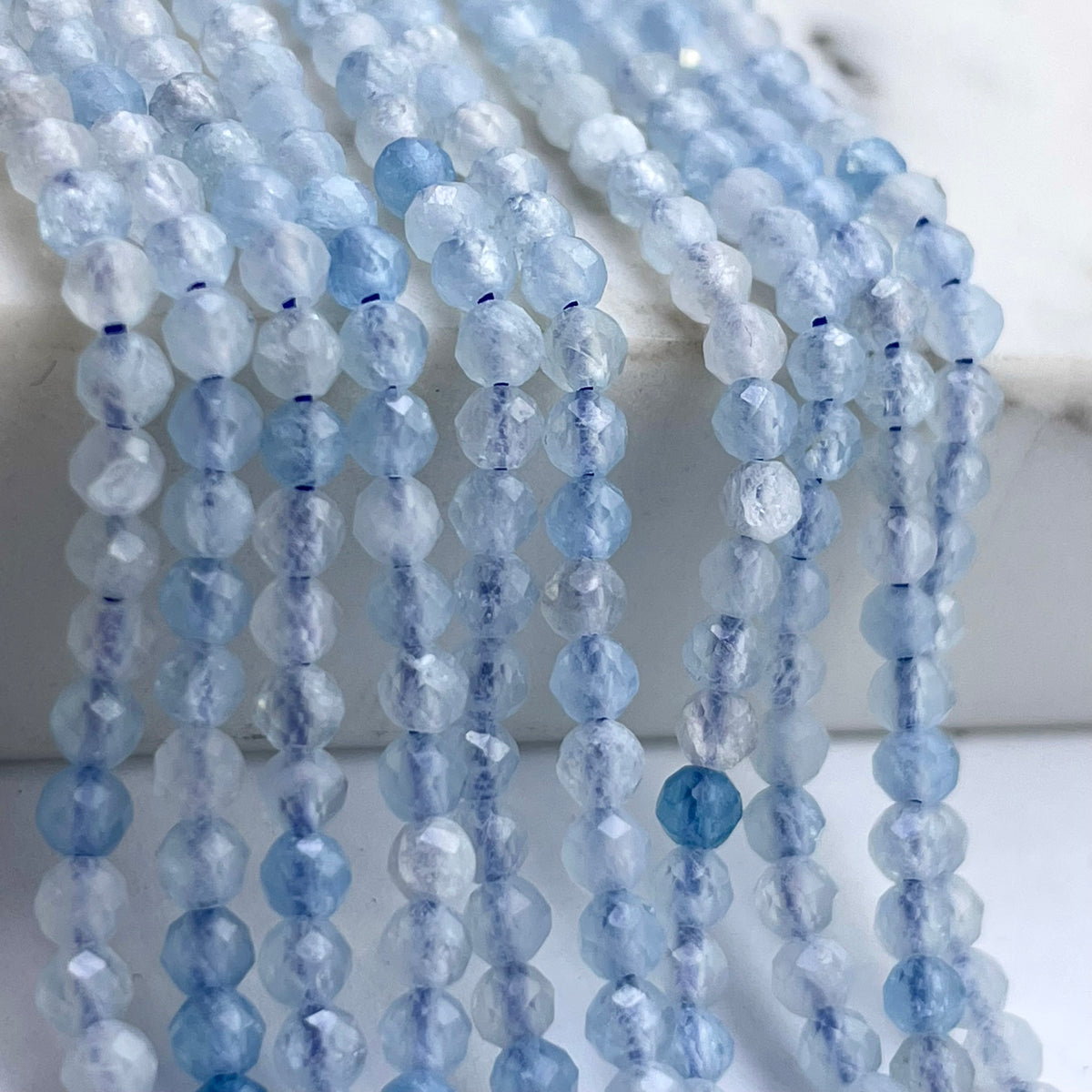 Faceted light blue aquamarine gemstone beads strung in multiple strands, draped over a white surface.