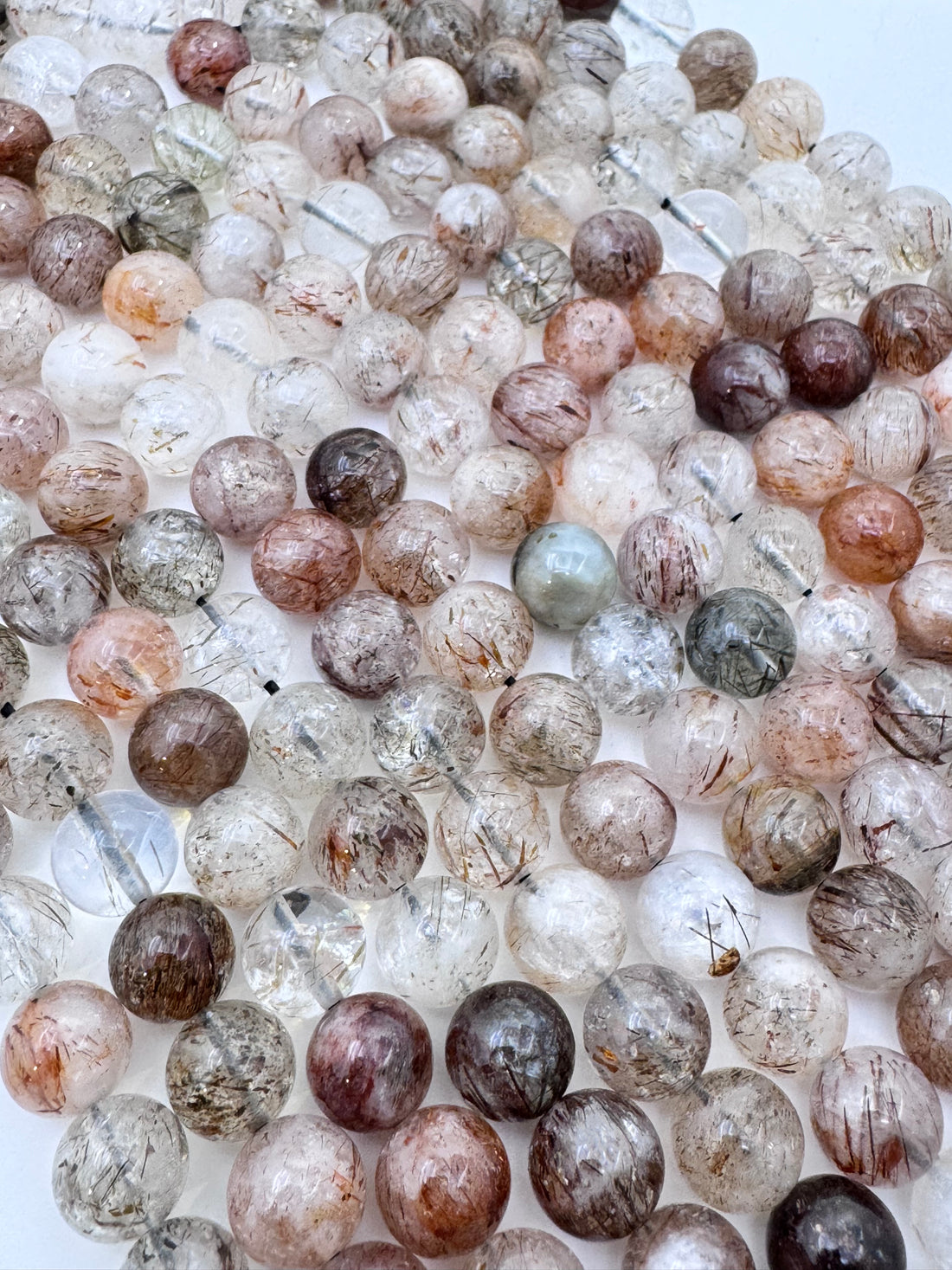 A close-up of a hand holding multiple strands of round black gold super seven quartz bead strands with visible inclusions and some iridescent reflections.