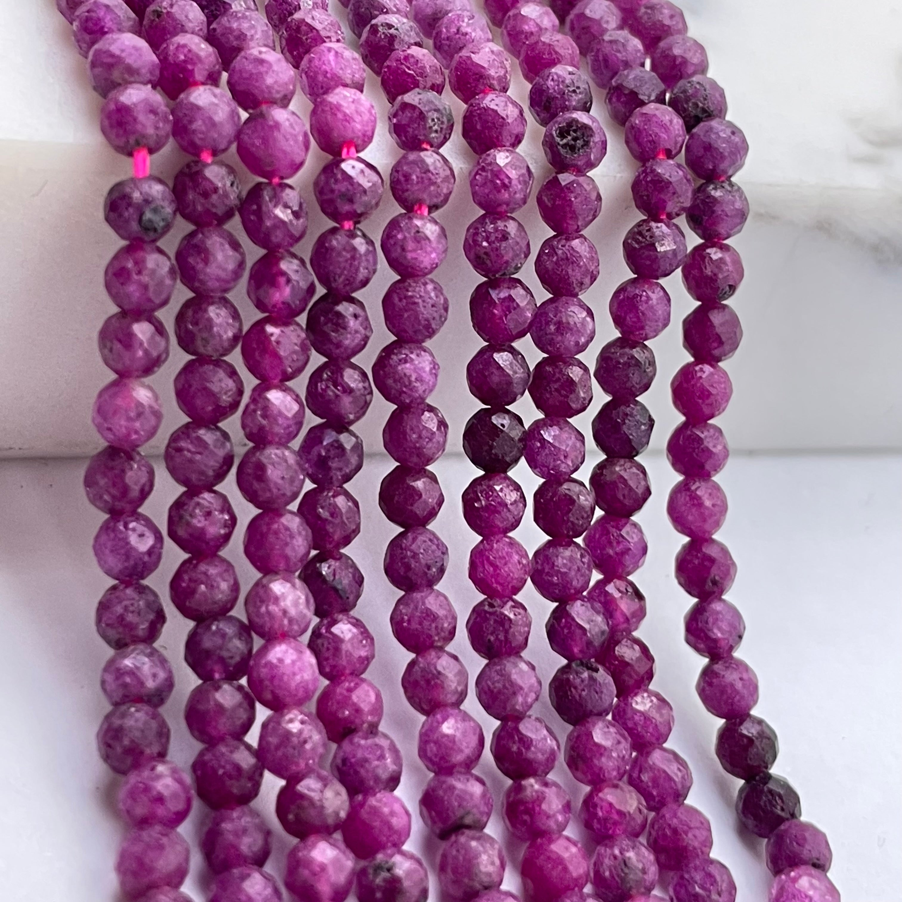Multiple strands of faceted purple ruby beads draped over a white marble surface.