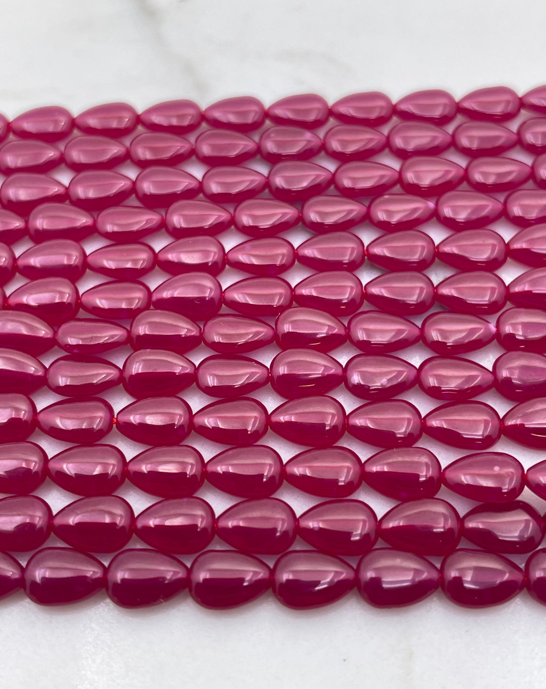 Close-up of glossy, teardrop-shaped lab ruby beads strung in multiple strands on a white surface, showing a vibrant magenta-pink color.