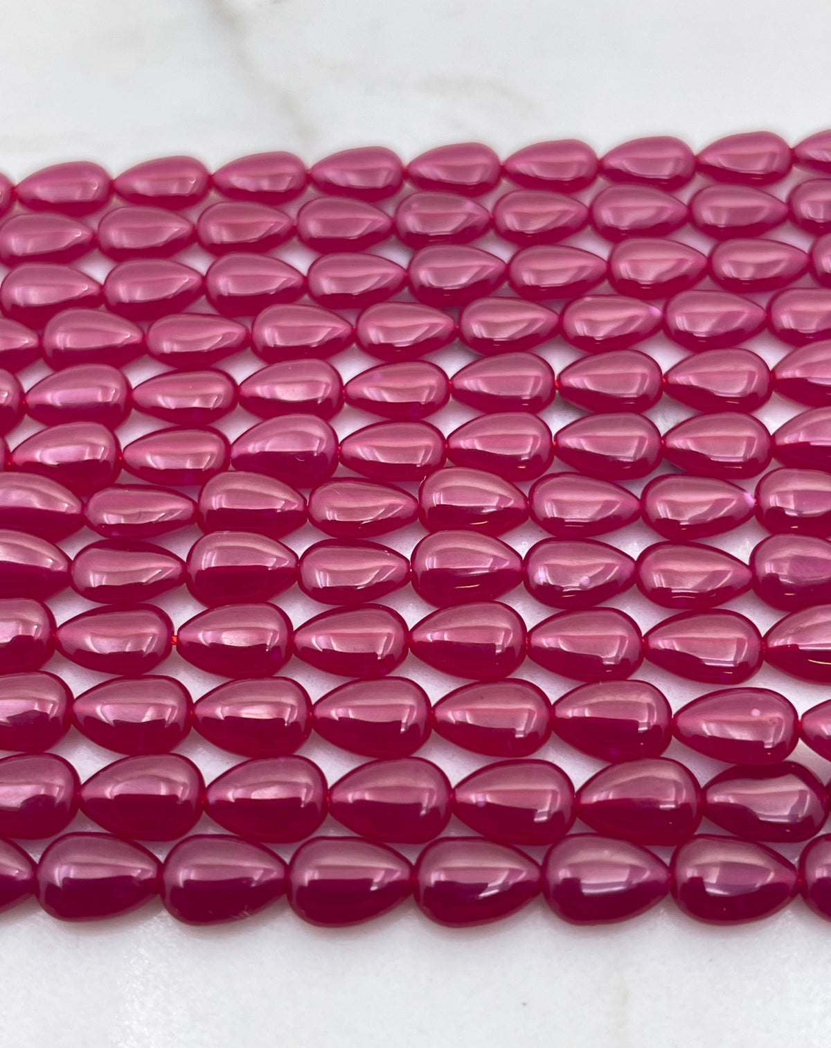 Multiple strands of glossy, teardrop-shaped lab ruby beads in a vibrant magenta pink color, arranged in parallel rows on a light marble surface.