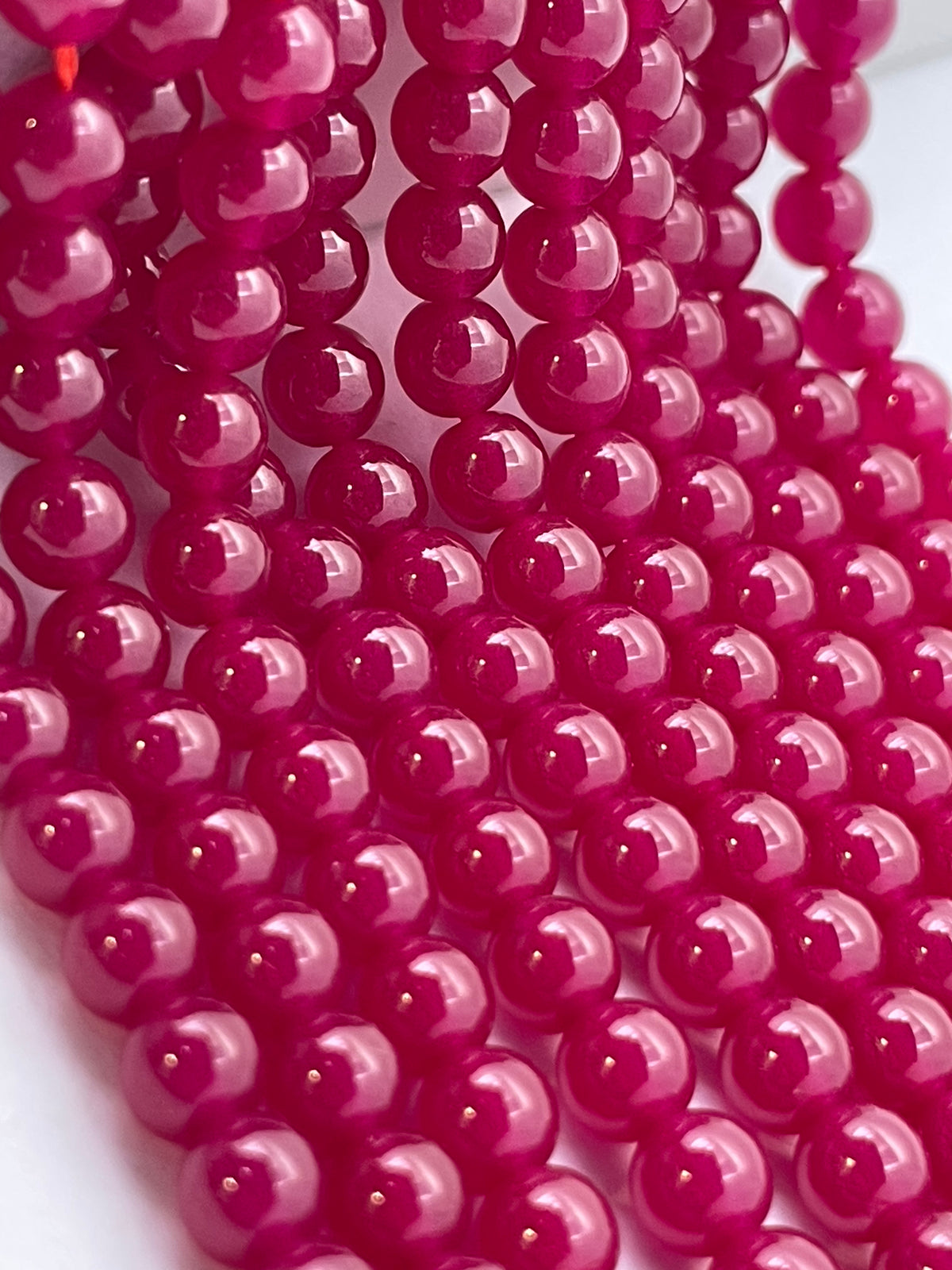 A close-up of glossy, round ruby beads in a vibrant magenta-pink color, arranged in parallel strands on a white background.