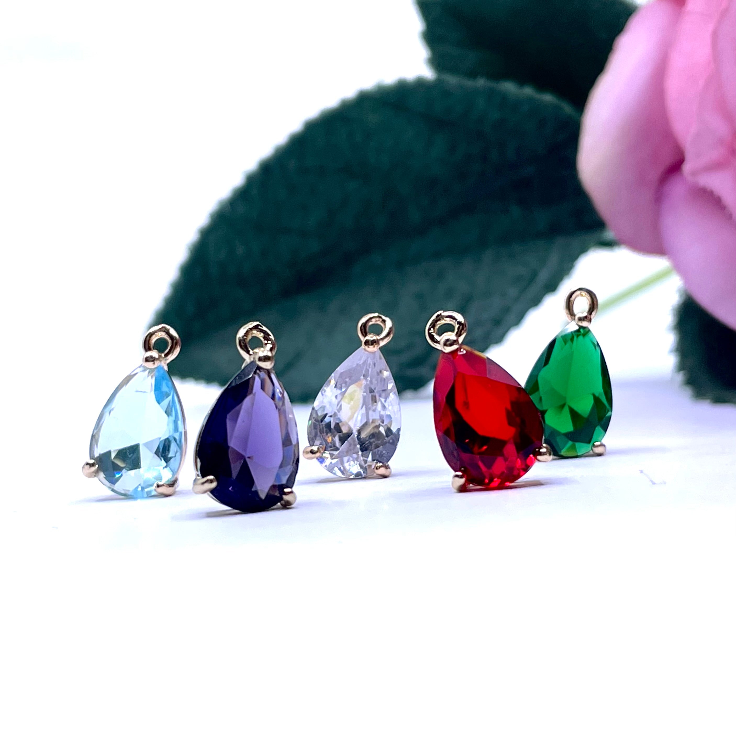Five teardrop-shaped faceted crystal pendants in light blue, dark purple, clear, red, and green, each with a gold-tone loop at the top, arranged on a white surface with blurred green leaves and a pink flower in the background.