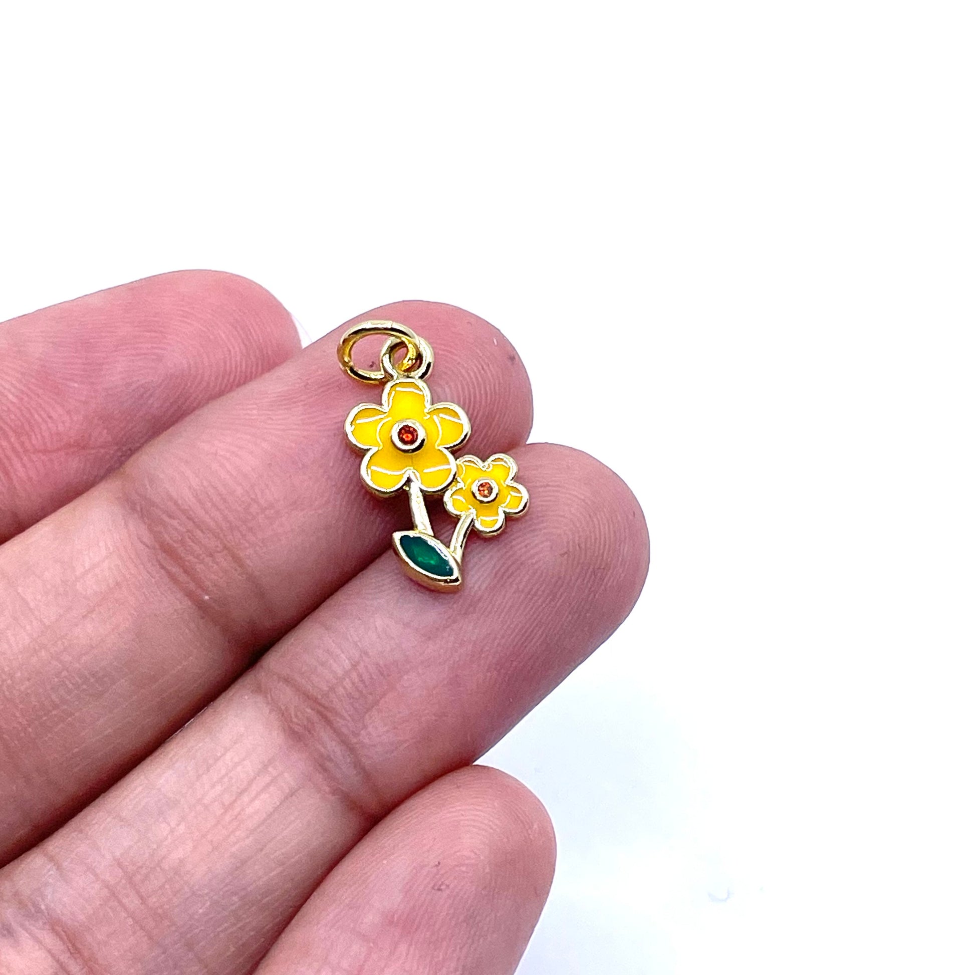 A small yellow flower charm with green leaves and gold-tone metal details, held between two fingers to show its size against a plain white background.