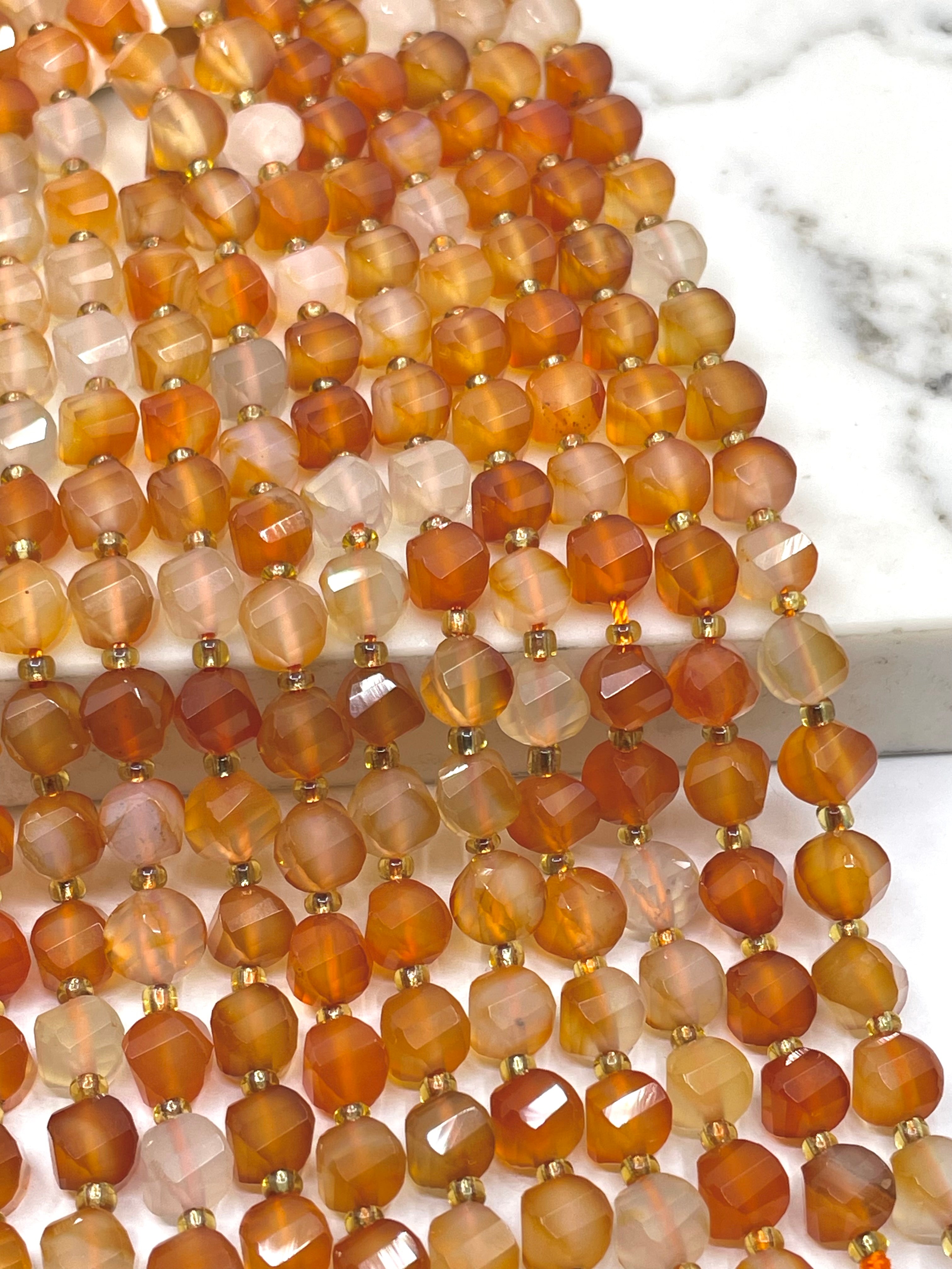 Multiple strands of faceted carnelian gemstone beads in warm amber, orange, brown, and translucent cream tones, separated by small gold spacer beads, arranged on a white marble surface.