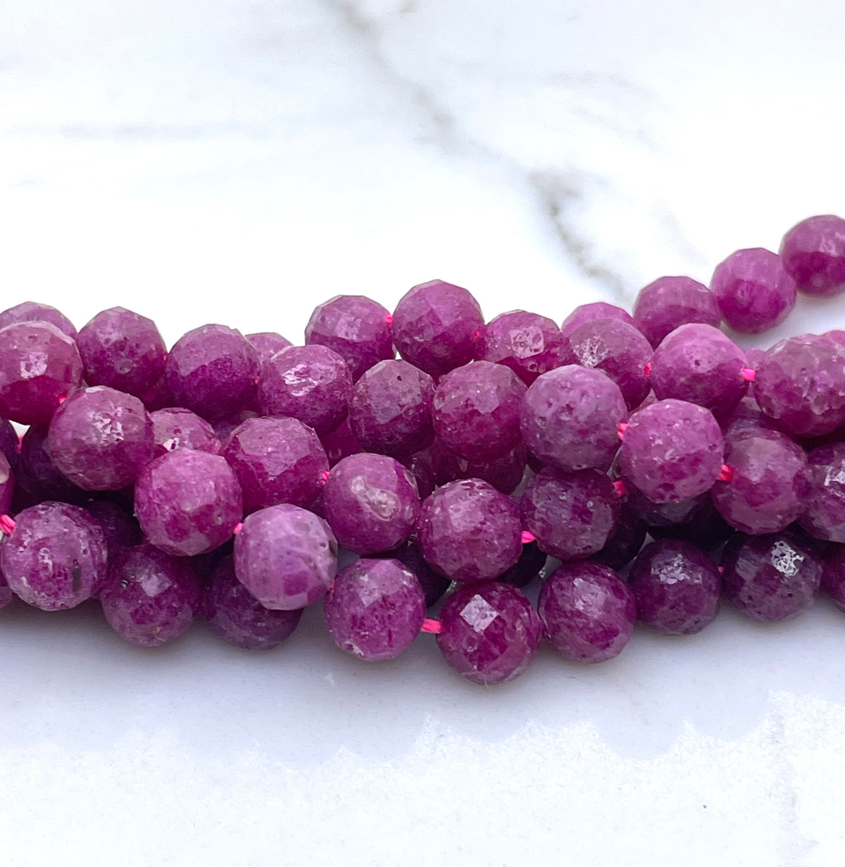 Faceted round ruby gemstone beads in a vibrant magenta-pink color, strung on a cord and arranged in a curve on a white marble surface.
