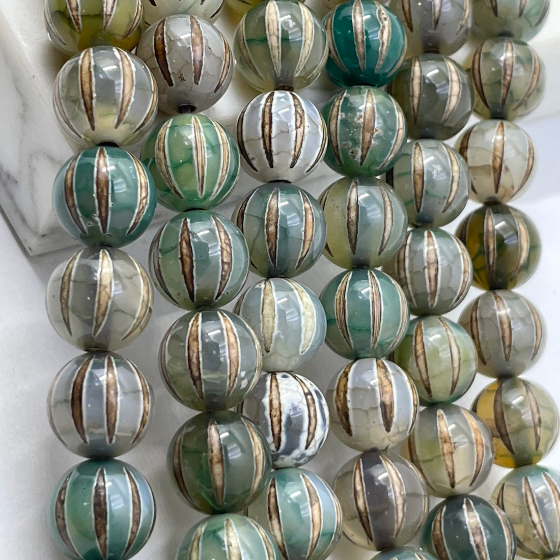 A close-up of round tibetan agate dzi beads strung in rows, featuring a marbled pattern of green, gray, white, and metallic gold vertical stripes.