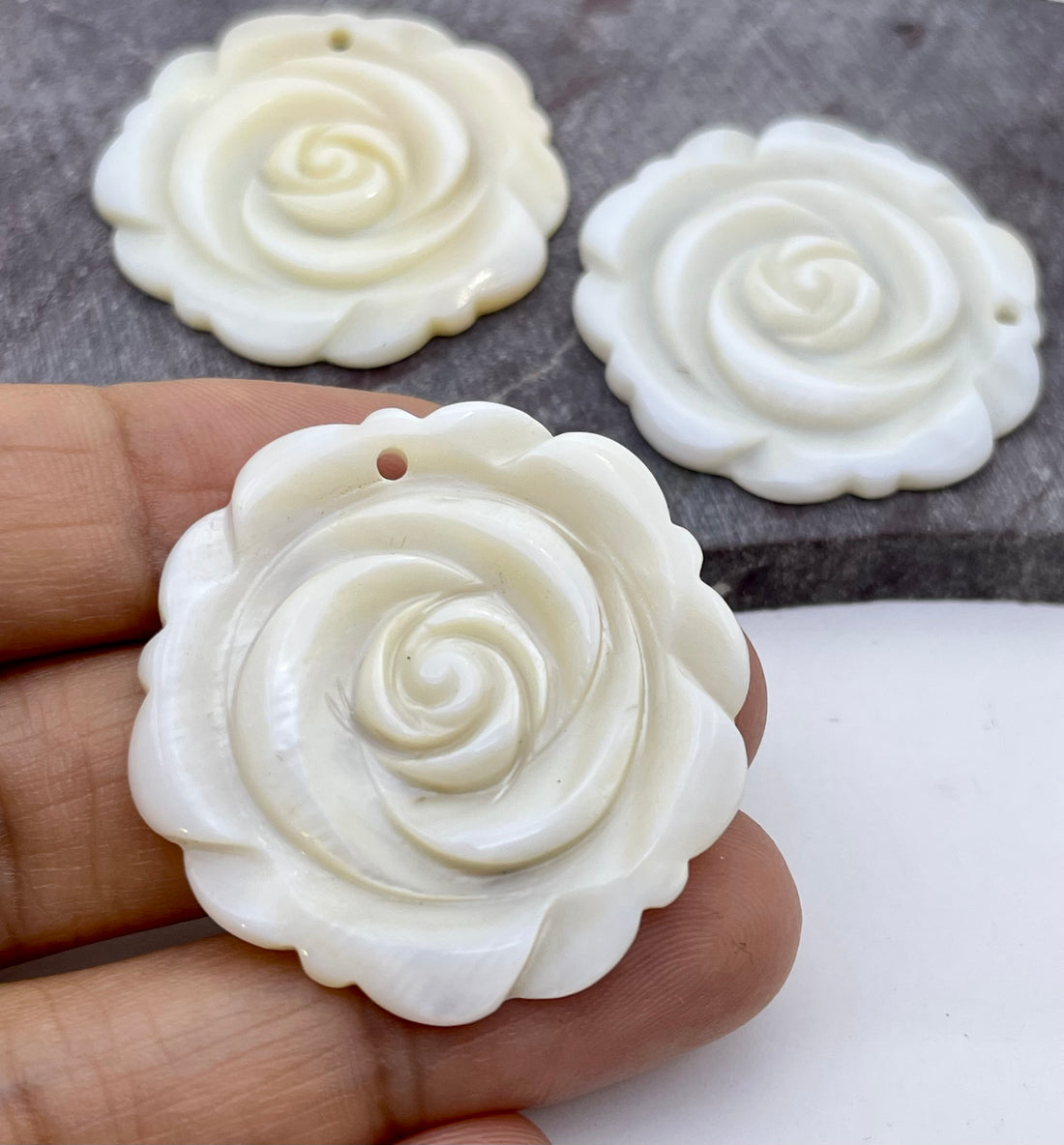 Three carved white rose-shaped jewelry pendants with a small hole at the top, shown with one held in a hand and two resting on a gray stone surface.