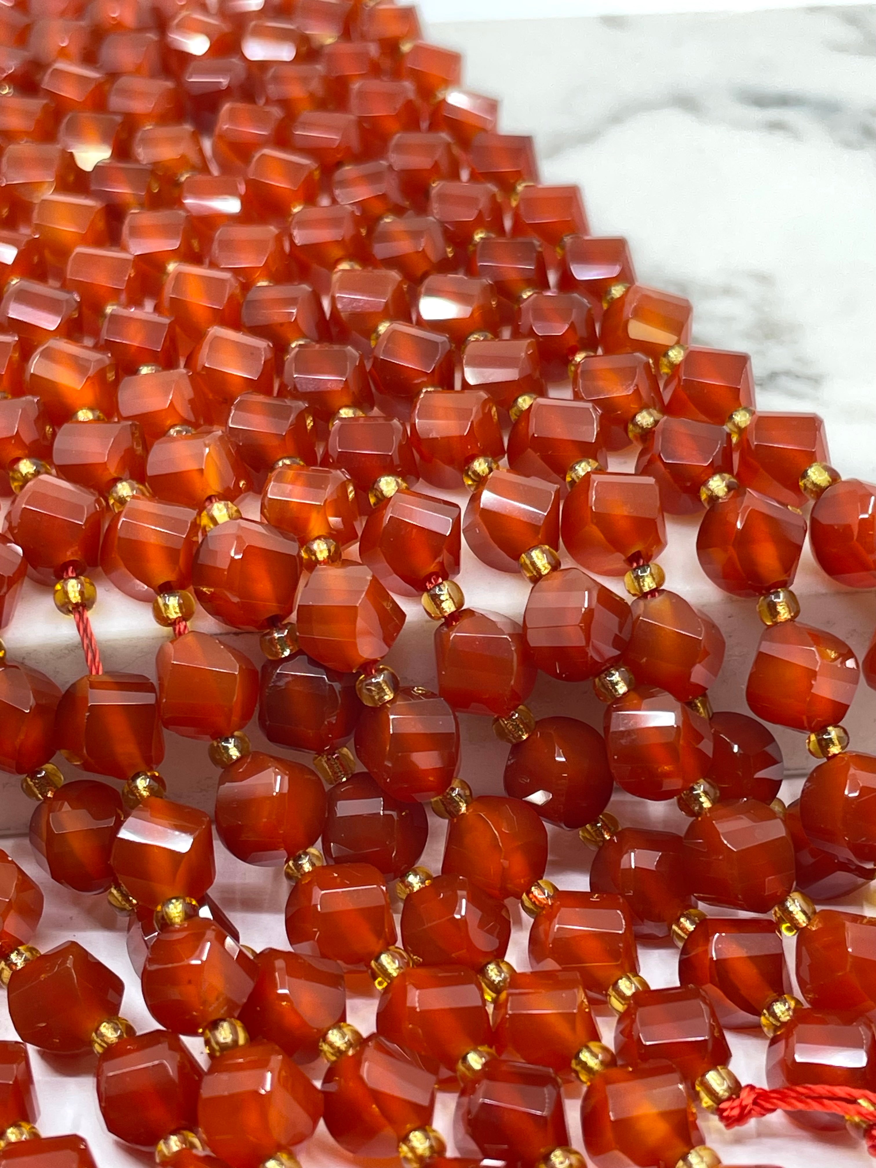 Faceted orange-red carnelian beads strung on cords with small gold spacer beads, photographed close up on a white marble-like surface.
