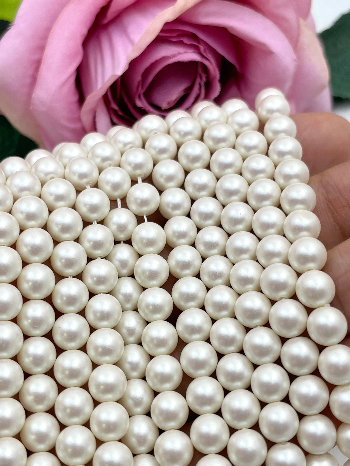 A close-up of strands of round, glossy ivory shell pearls draped over a hand, with a soft pink rose in the background.