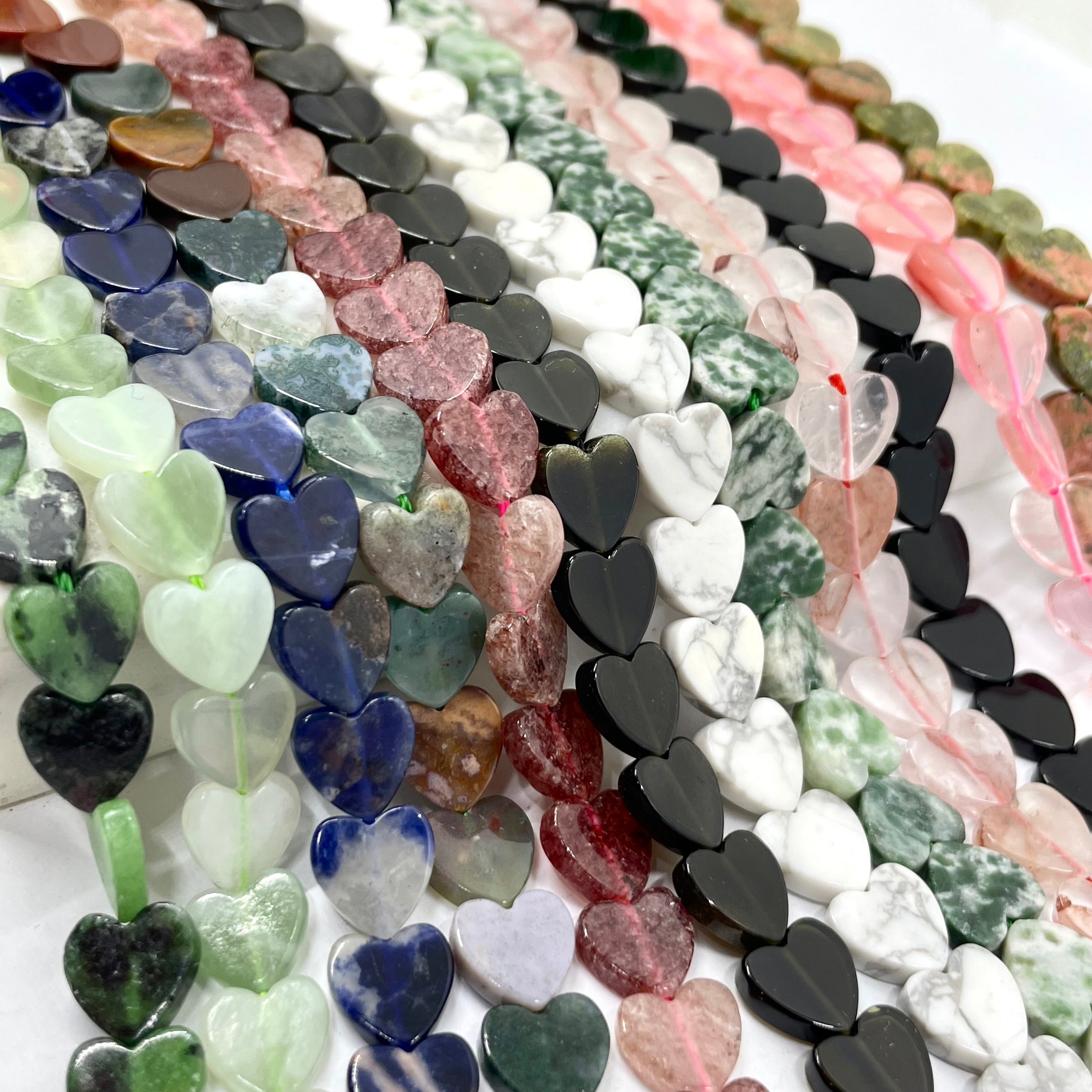 Multiple strands of heart-shaped gemstone beads arranged diagonally, showing a variety of colors and natural stone patterns including pink, green, blue, white, and black.