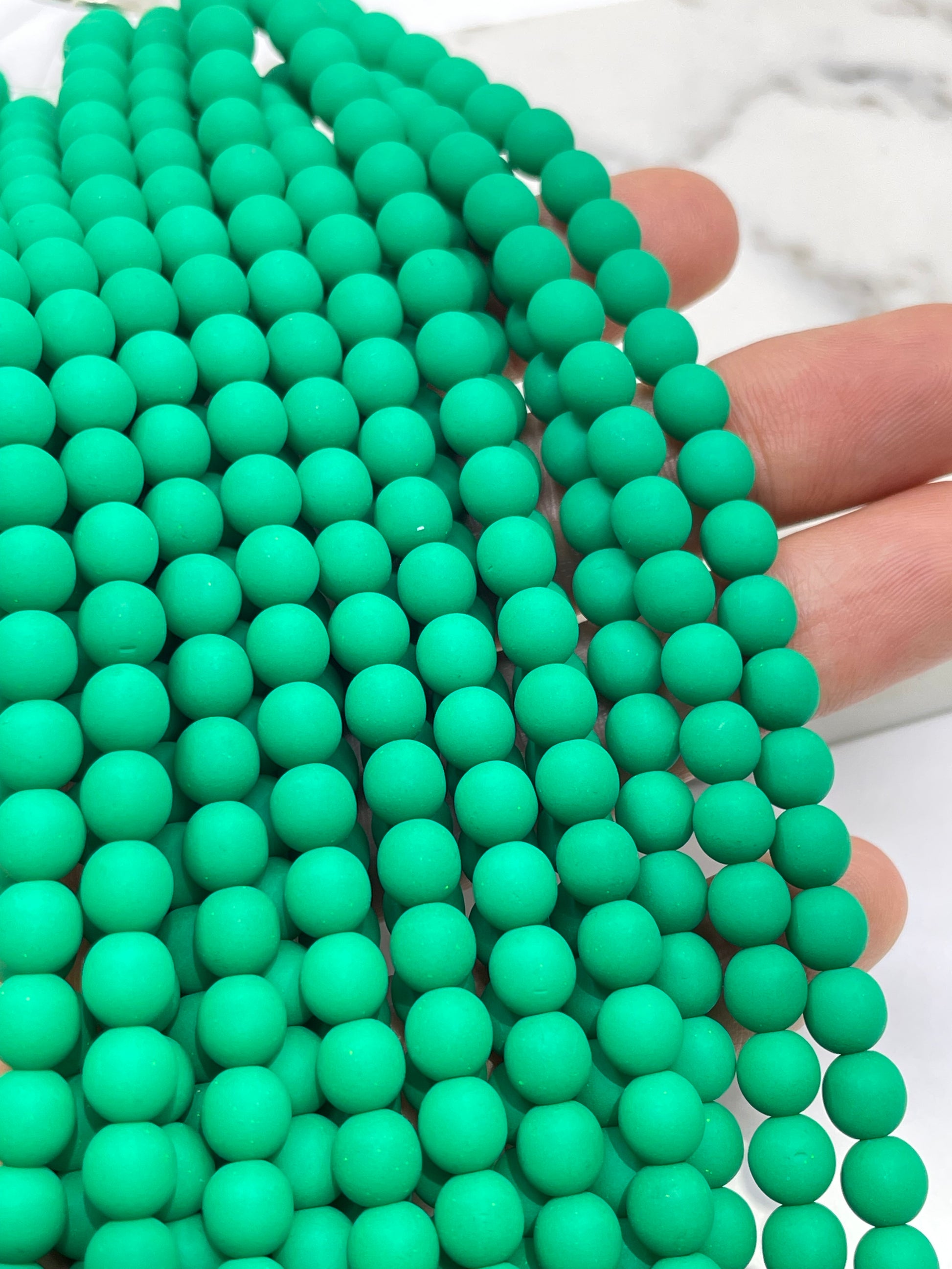 A close-up of a hand holding multiple strands of matte mint green round beads, with fingers visible behind the beads against a light marble-like background.