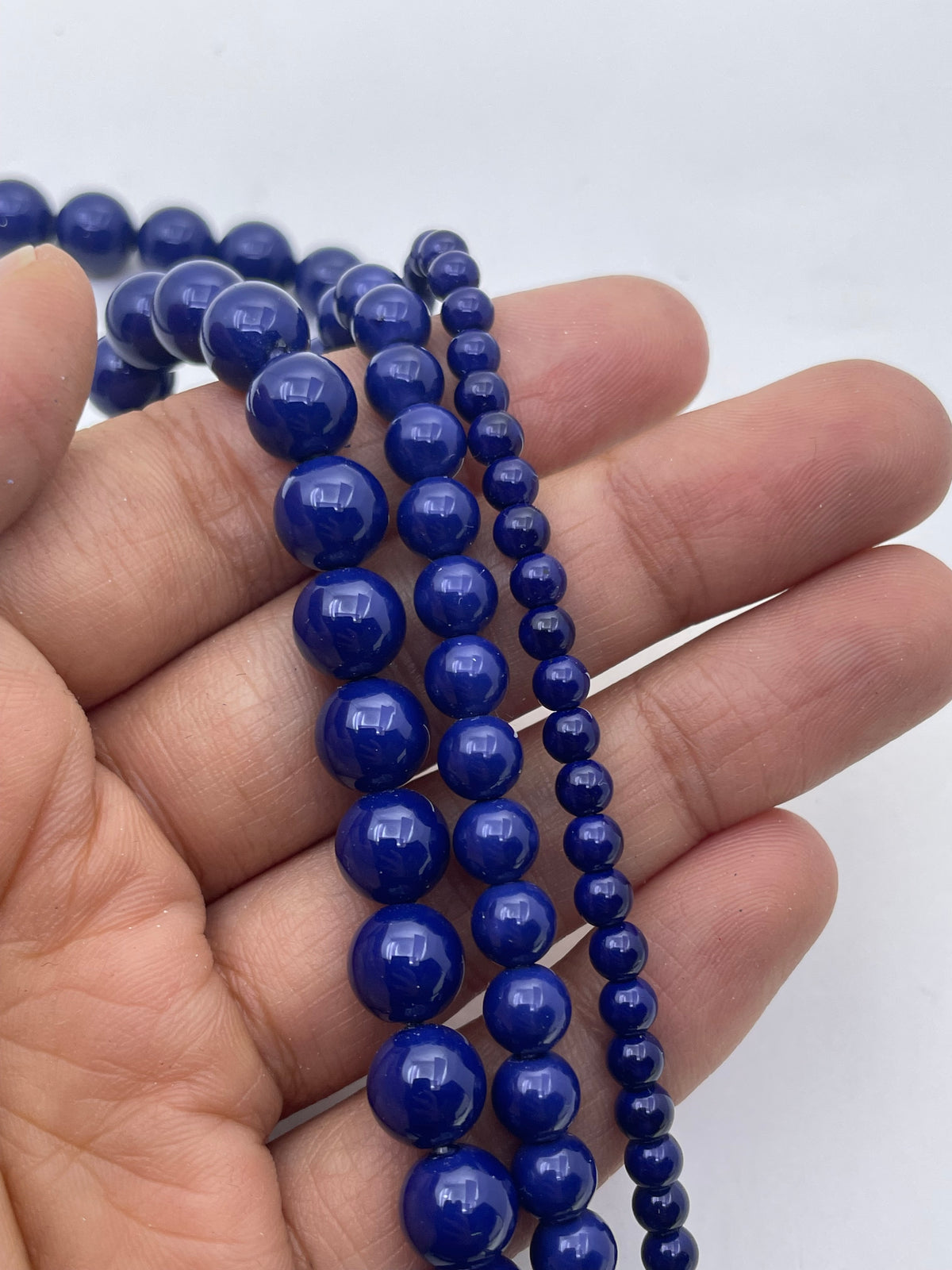 A close-up of a hand holding three strands of polished cobalt blue shell pearl beads, with the strands curving downward against a plain white background.