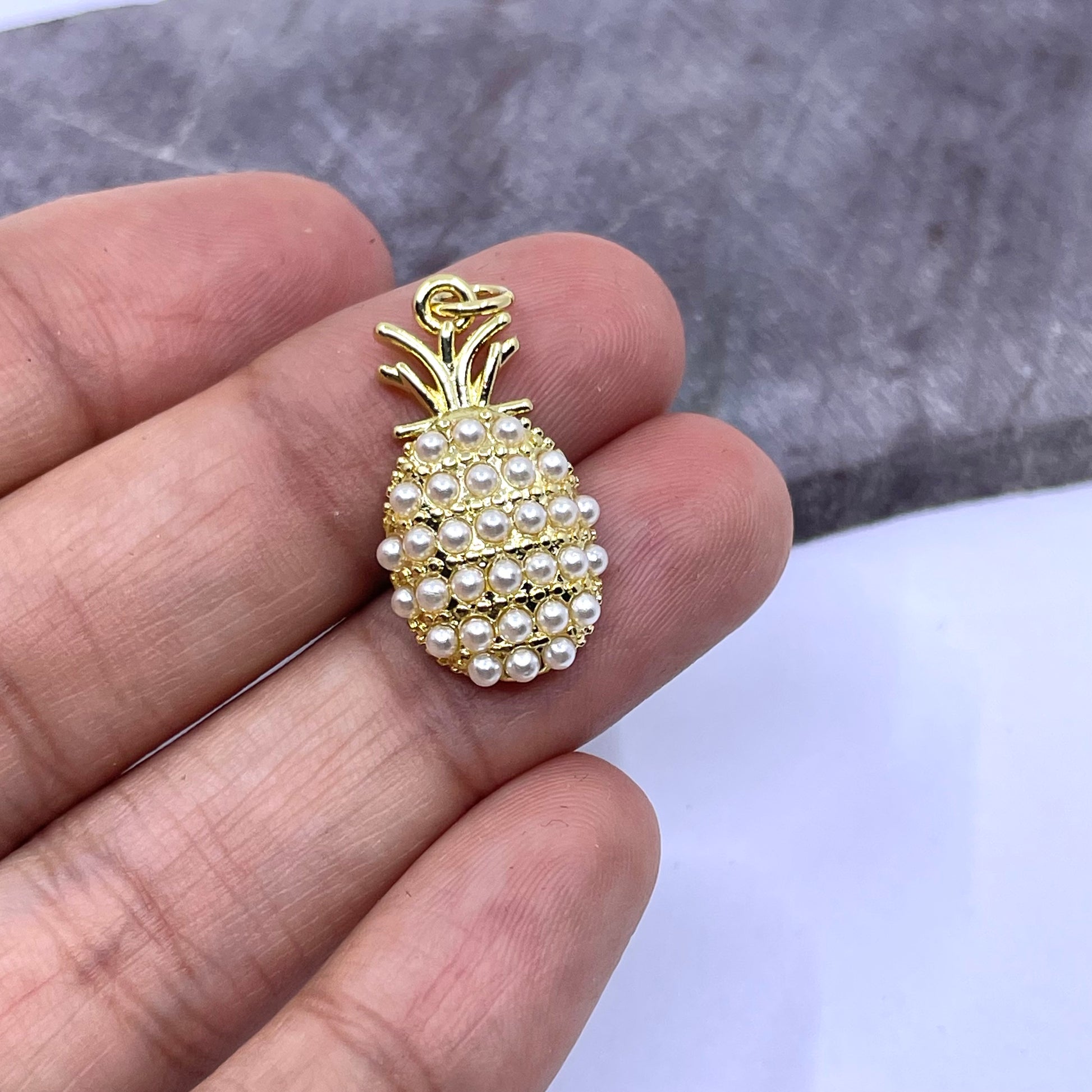 A small pineapple-shaped jewelry charm or pendant made of gold-toned metal and white faux pearls, held between fingers for scale against a gray and white background.