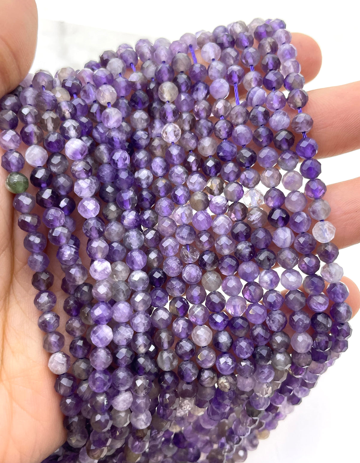 Faceted purple amethyst gemstone beads held in a hand, showing multiple strands of round beads with natural color variations from light lavender to deep violet.