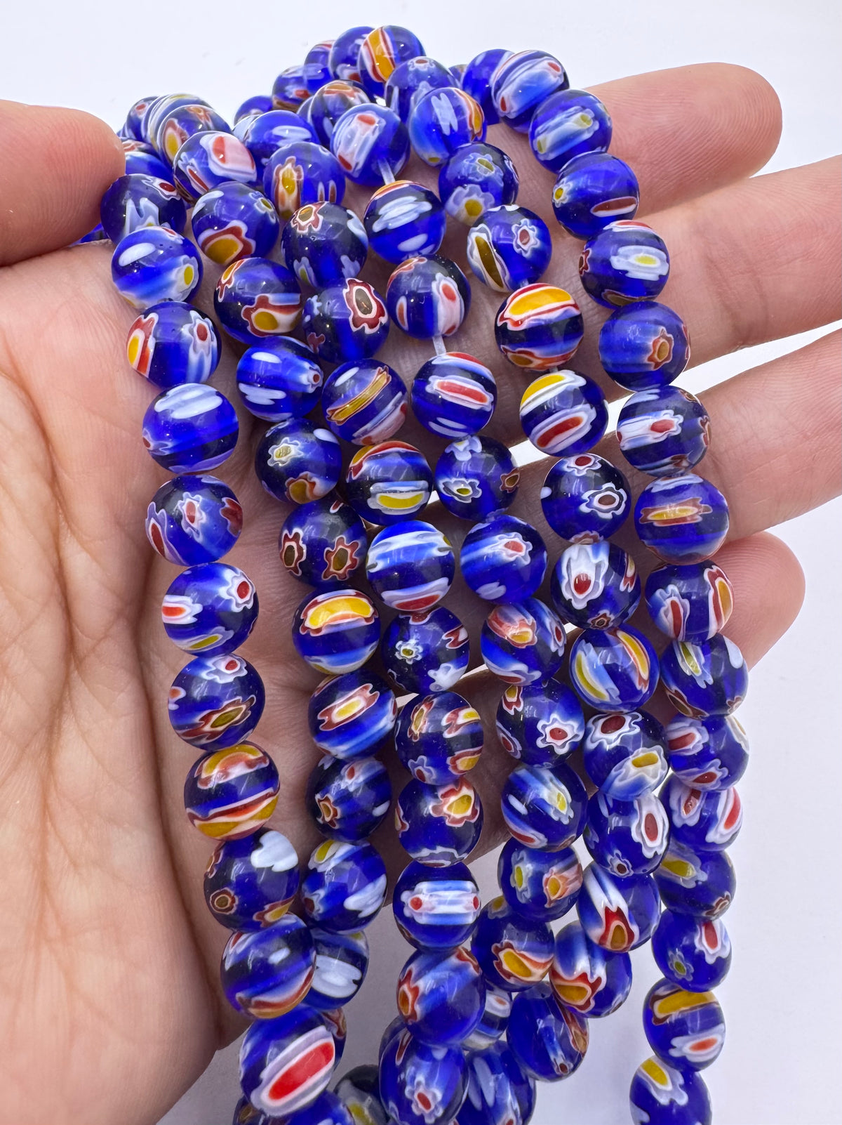 A hand holding a strand of round murano glass beads, featuring a deep cobalt blue base with intricate swirls of white, orange, yellow, and red.