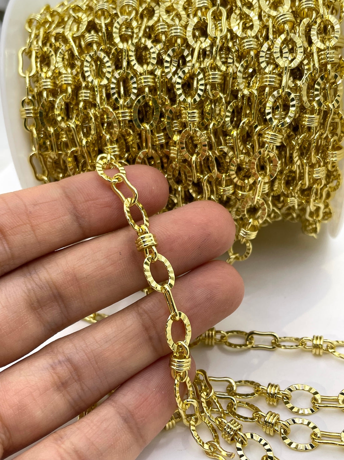 A close-up of a gold-colored chain link accessory, showing a spool of the chain in the background and several individual links draped in the foreground on a white surface.