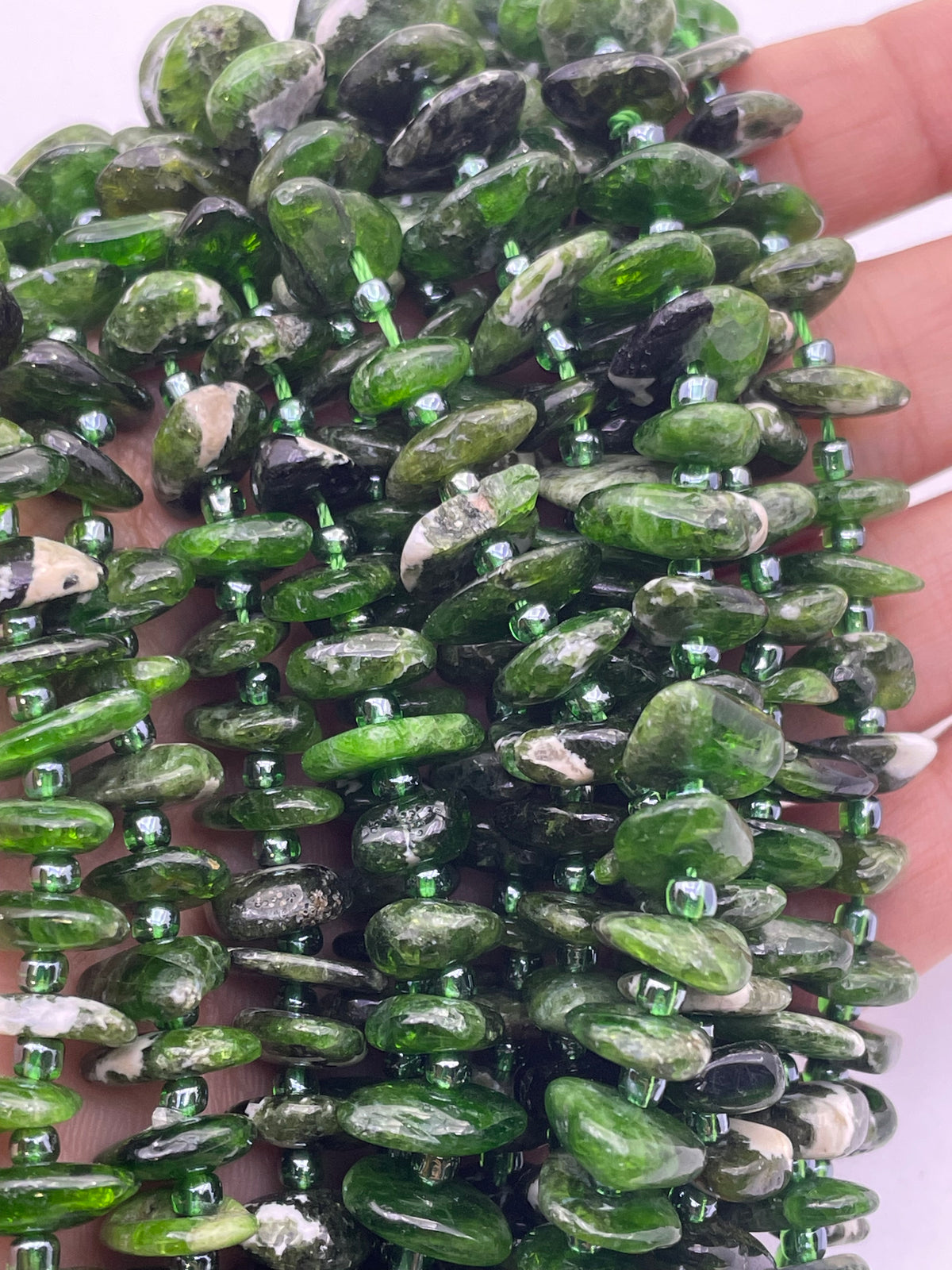 A close-up of a hand holding a strand of polished diopside stone beads, with some darker inclusions and small metallic spacer beads.