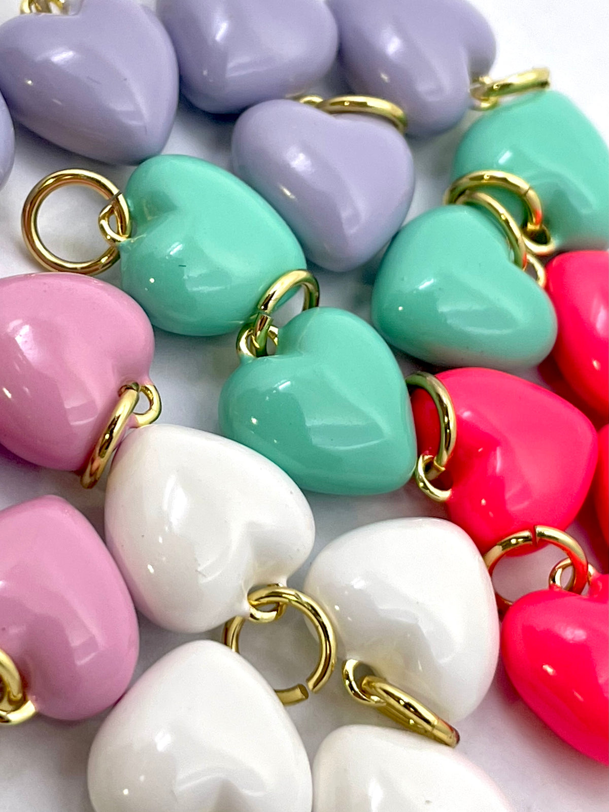 A close-up of glossy heart-shaped charms in pastel lavender, mint green, pink, white, and bright pink, each attached with a gold ring clasp.