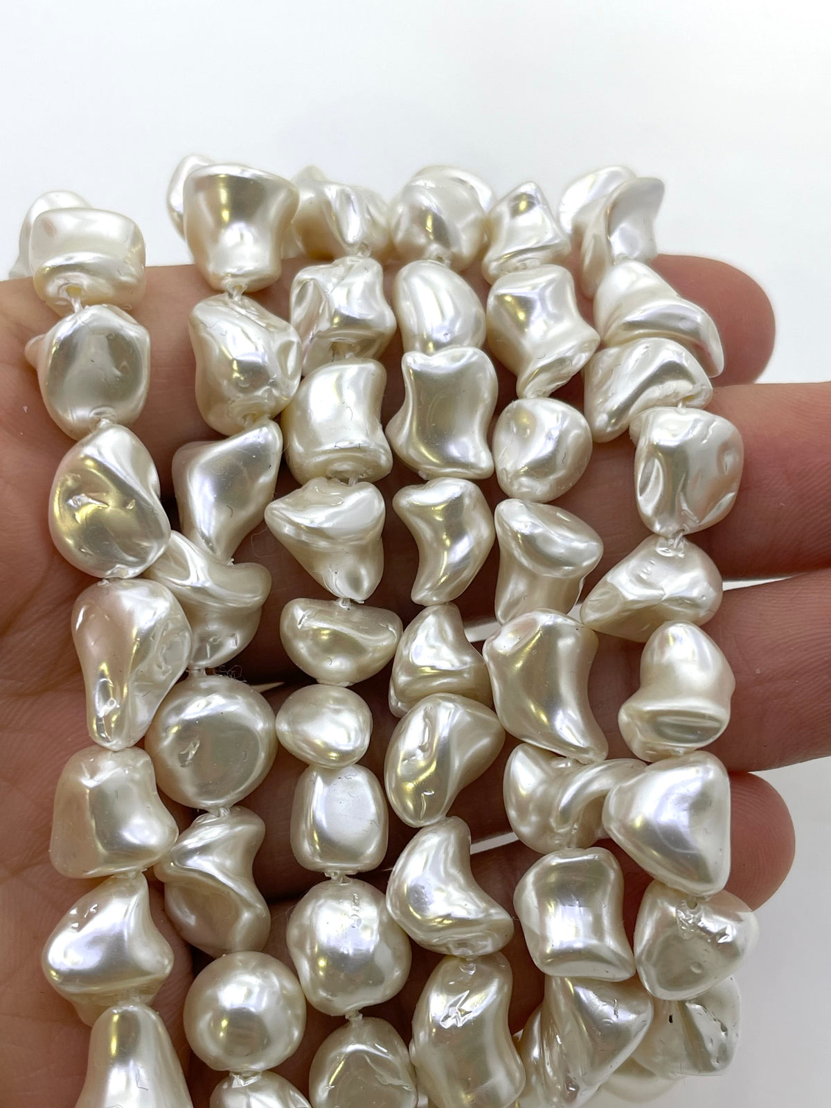 A close-up of irregular, lustrous white baroque shell pearl beads held in a hand against a plain white background.