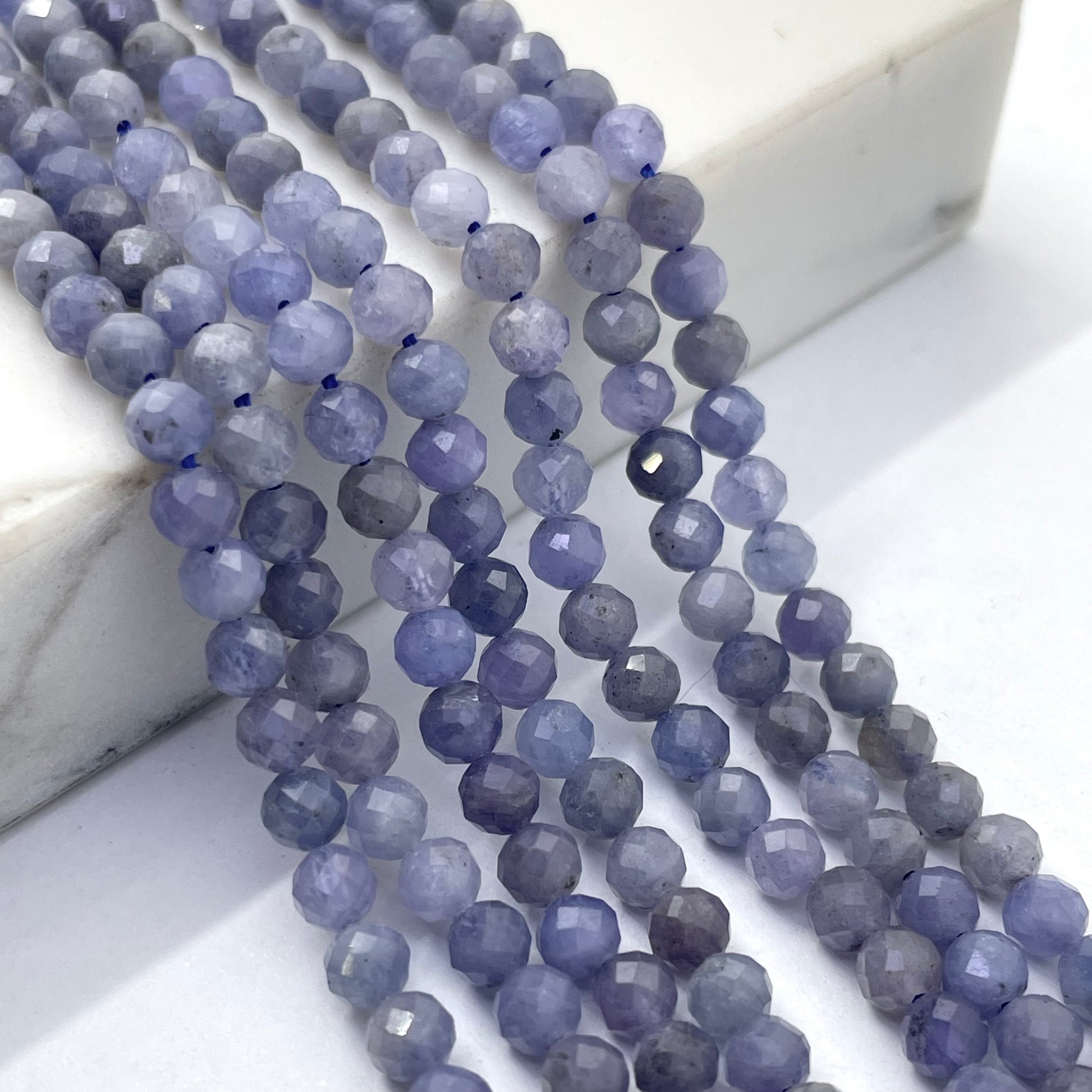 Faceted round tanzanite beads in a dusty lavender-blue color arranged in parallel strands on a white marble block and white surface.