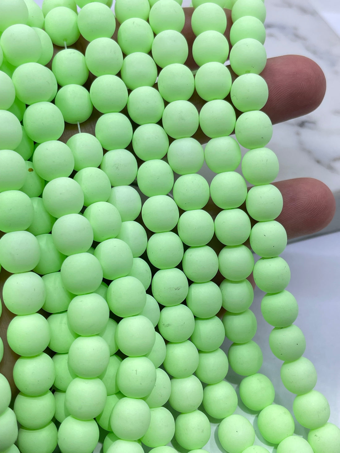 A close-up of many pale green grape-like beads called velvet beads or pom-pom balls arranged in rows, with a small white marble-like surface visible in the upper left corner.