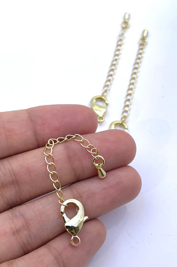 A close-up of a hand holding a gold-tone lobster clasp and extension chain, with two similar gold chain and clasp pieces placed on a white surface in the background.