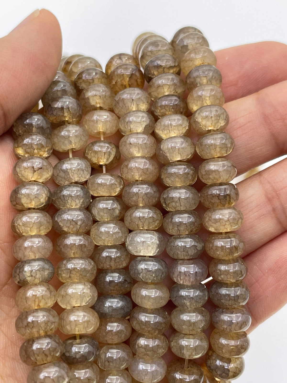 A close-up of a hand holding several strands of smooth donut translucent brownish-gray glass beads on a plain white background.