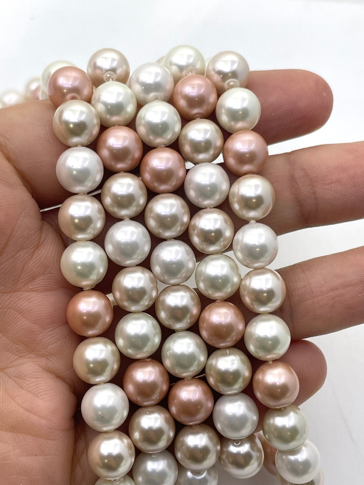 A hand holding a strand of shell pearl beads in three colors: white, soft pink, and champagne gold, shown against a plain white background.
