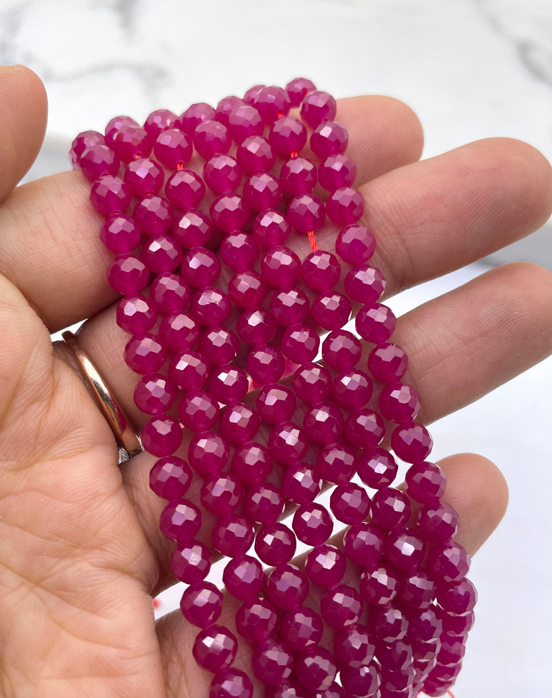 Faceted round ruby beads in a vibrant magenta-pink color, arranged in parallel strands on a white marble-like surface.
