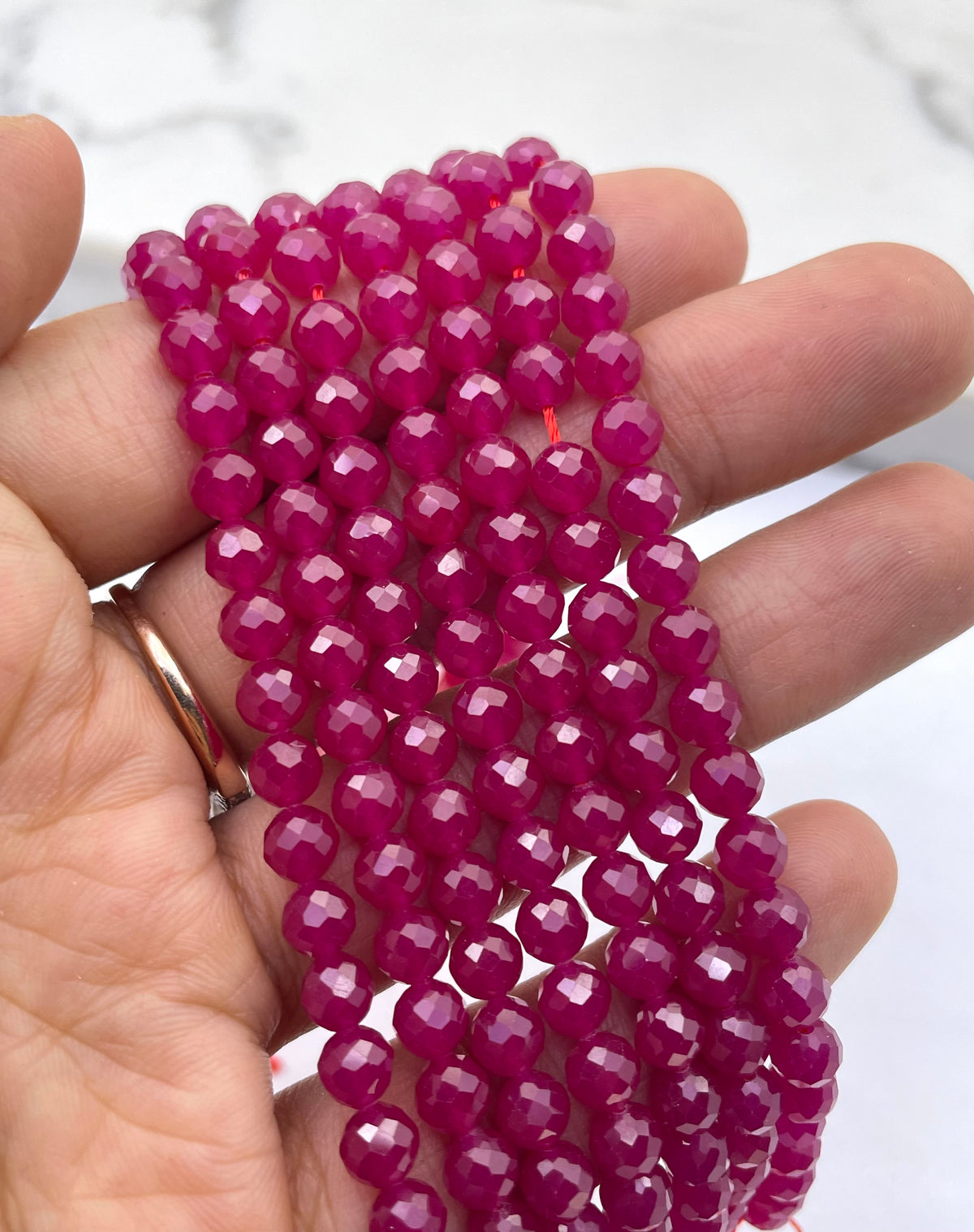 A hand holding a bundle of faceted ruby magenta beads, likely for jewelry making, against a light marble background.
