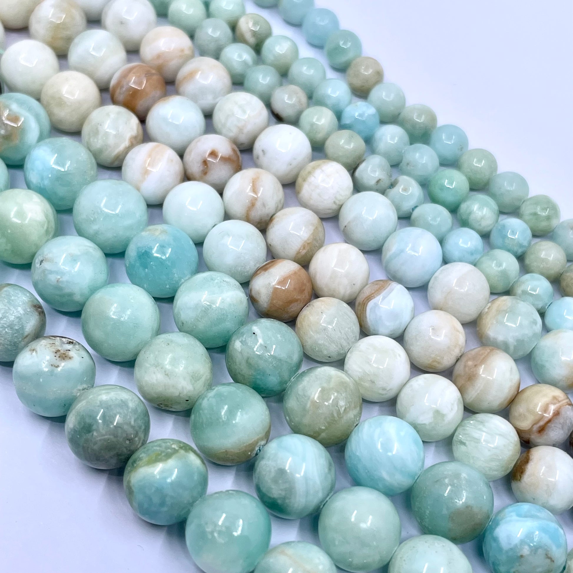 A close-up of multiple strands of polished round hemimorphite gemstone beads in light blue-green, white, and soft beige with natural marbling, arranged on a pale background.