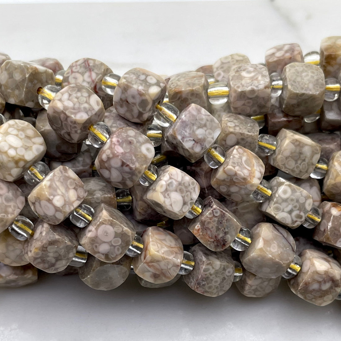 Close-up of a strand of faceted gemstone beads in a mottled beige, brown, and white pattern, threaded with small clear glass and metallic gold spacer beads on a white marble surface.
