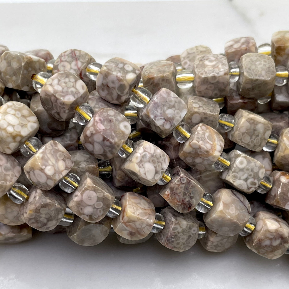 Close-up of a strand of faceted gemstone beads in a mottled beige, brown, and white pattern, threaded with small clear glass and metallic gold spacer beads on a white marble surface.