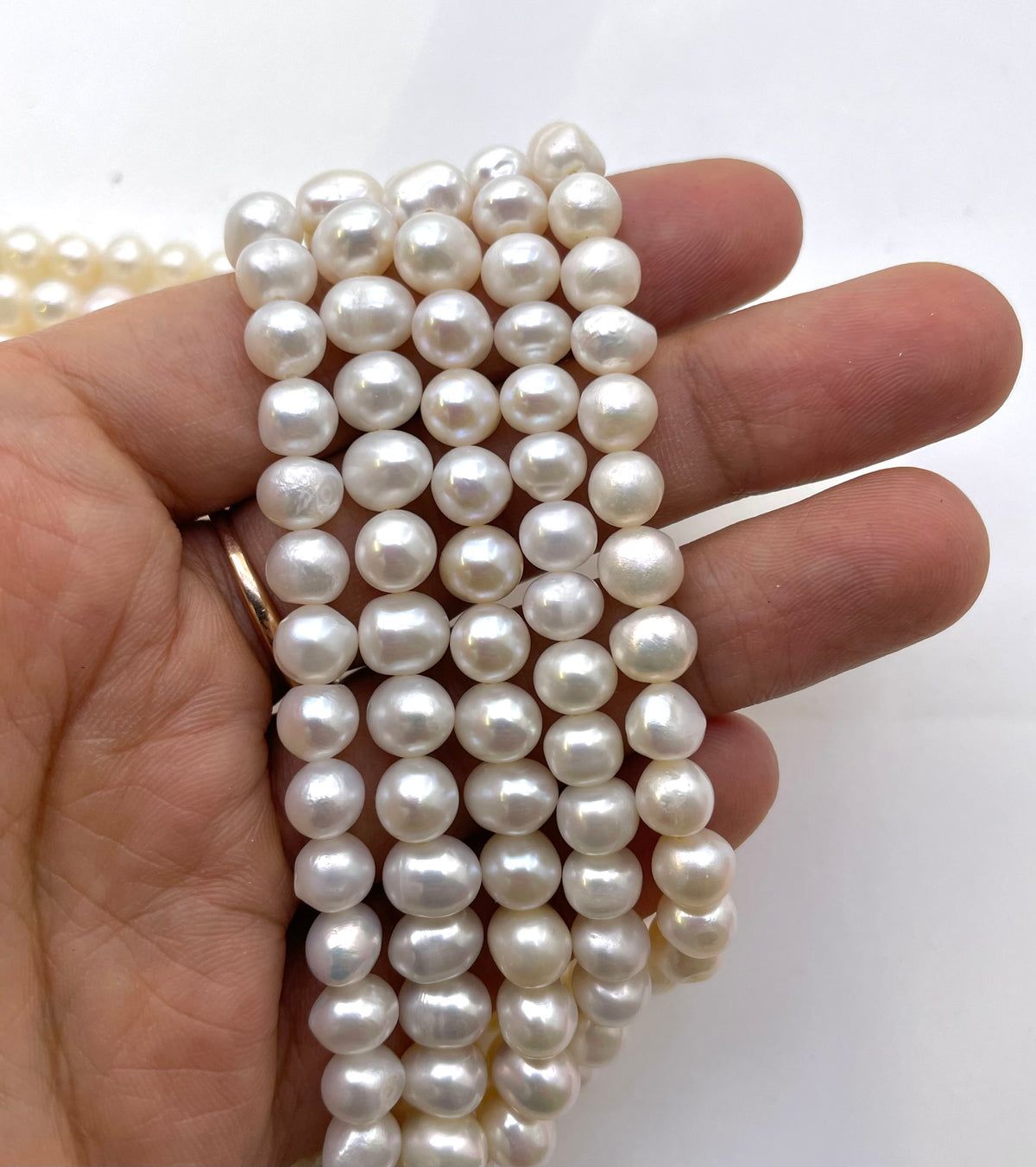 A close-up of a hand holding a bundle of strands of white potato freshwater pearls, with more pearls visible in the background.