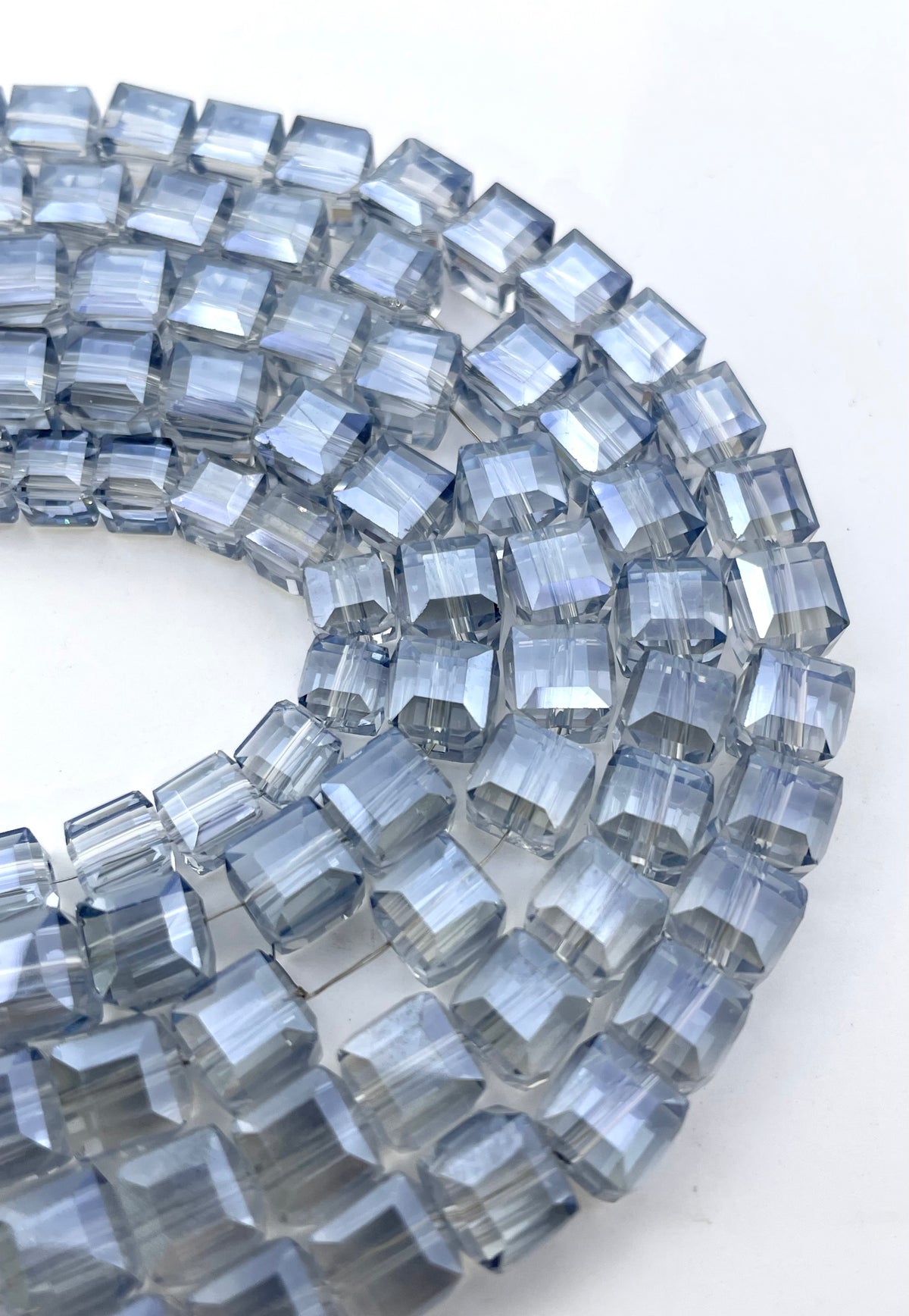 A close-up of faceted cube glass beads in a smoky translucent gray-blue color, arranged in a curved line on a plain white background.