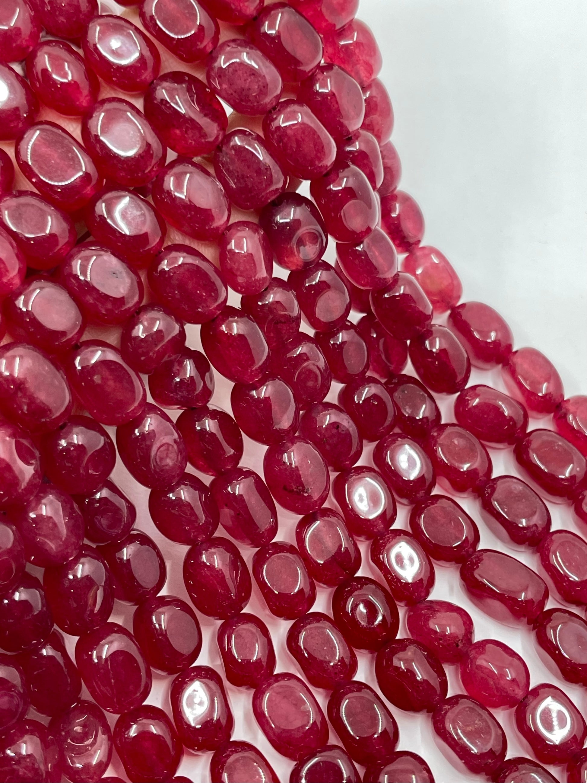 Multiple strands of polished oval jade gemstone beads in a deep raspberry pink color, arranged diagonally on a plain white background.