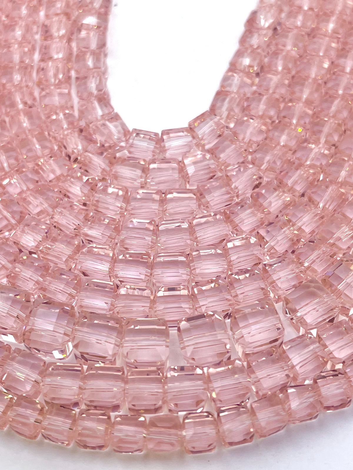 A close-up of a strand of faceted, cube rose pink glass beads arranged in a curved, layered pattern against a white background.