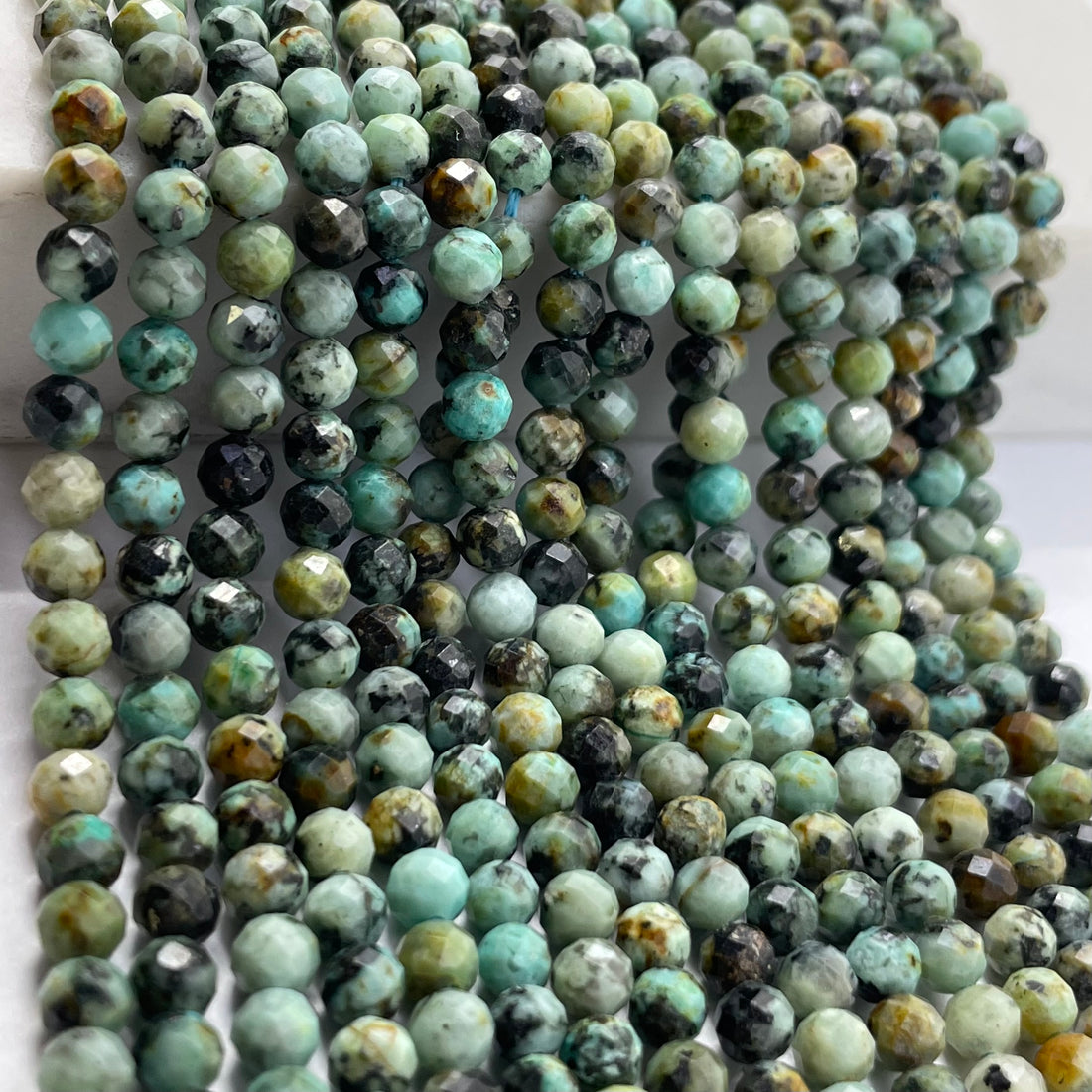 Multiple strands of faceted round african turquoise beads in a marbled mix of turquoise, green, black, and beige tones, draped over a white surface.