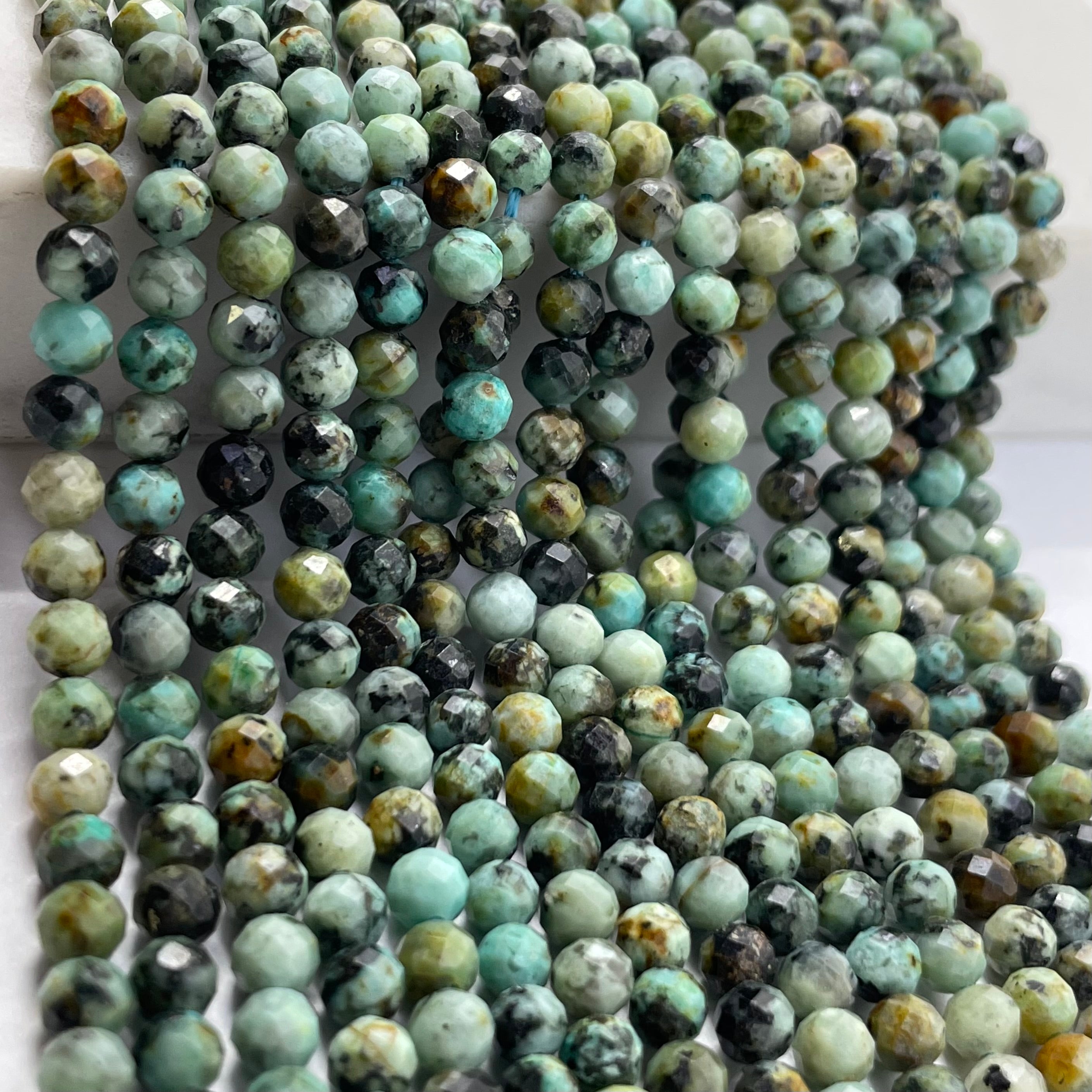Multiple strands of faceted round african turquoise beads in a marbled mix of turquoise, green, black, and beige tones, draped over a white surface.