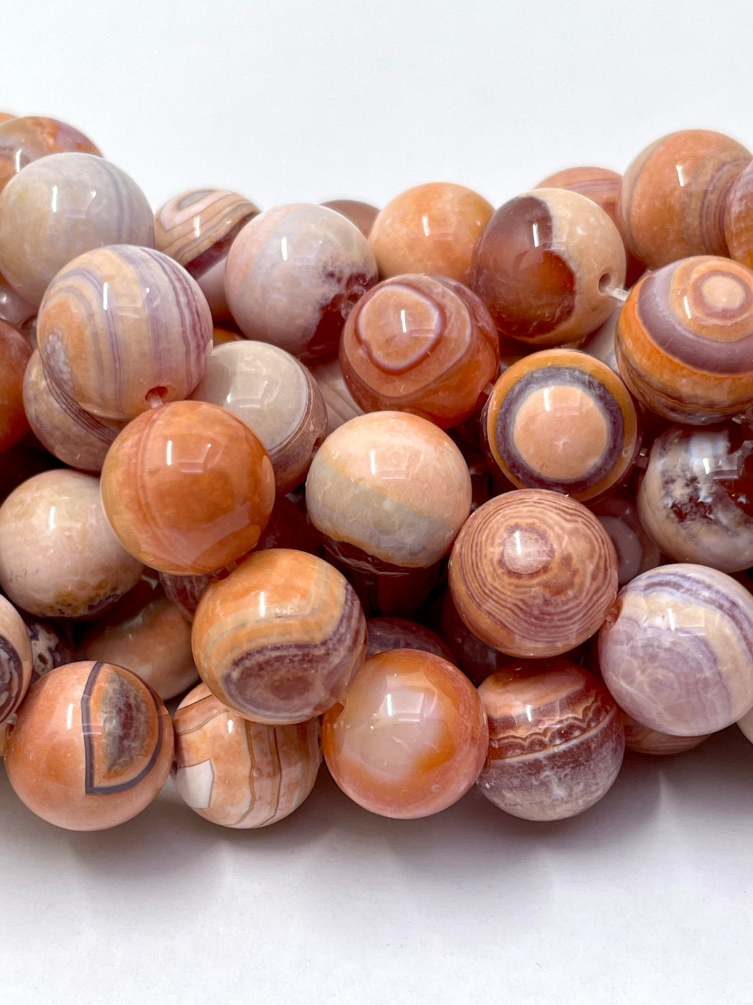 A close-up of polished round gobi agate gemstone beads in earthy orange, peach, cream, and lavender-brown banding patterns, arranged on a plain white background.