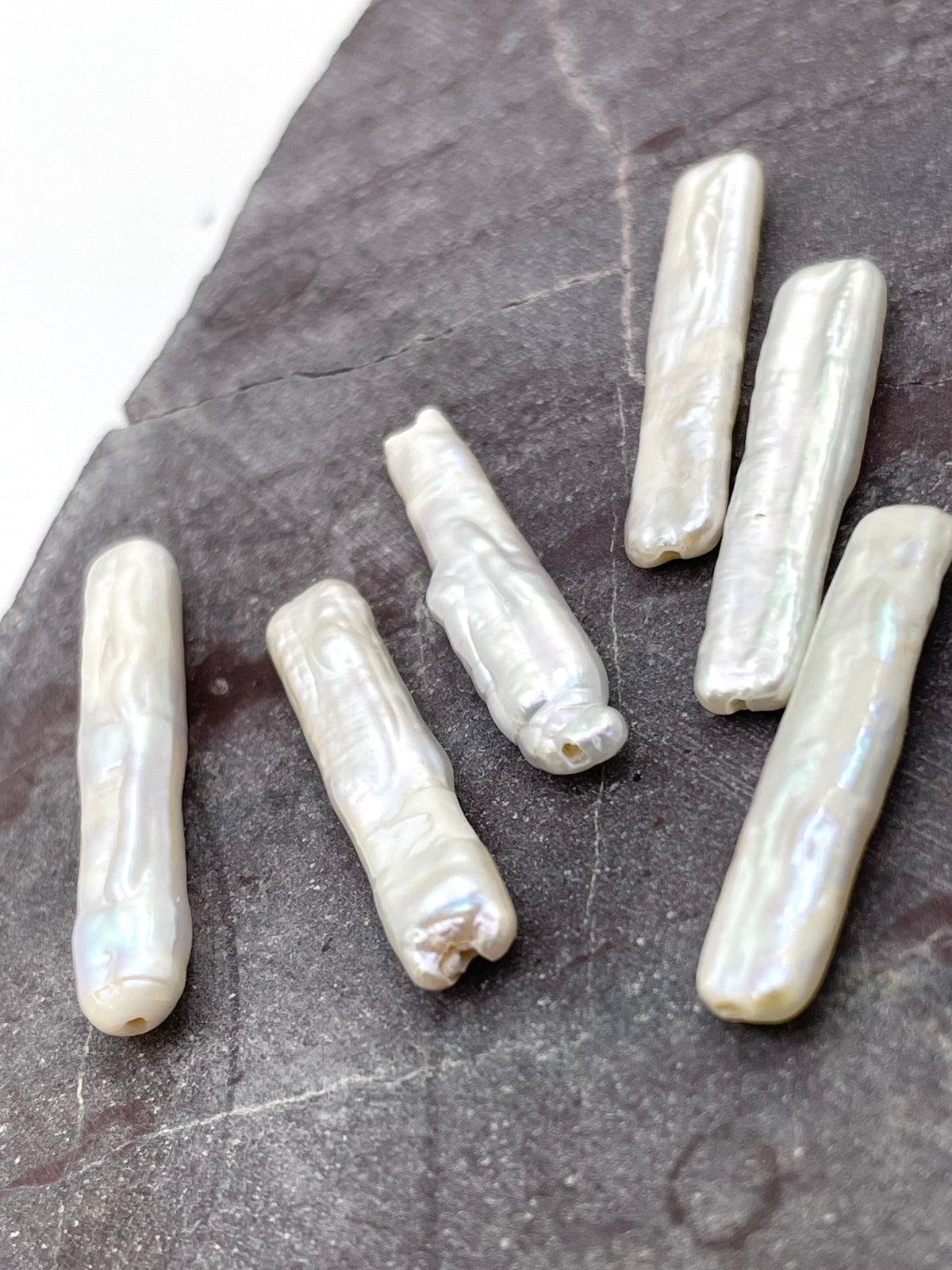 Six irregular baroque-style white pearl beads with natural luster, arranged on a dark textured slate surface.